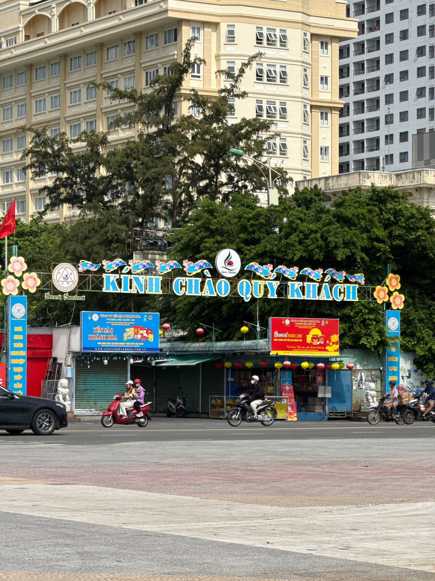 This is a street-level, eye-level, medium shot of a busy intersection in Nha Trang, Vietnam, on a sunny day. In the foreground, a paved intersection with a red and white patterned crossing is visible. On the left side of the frame, a black car is parked. Two motorcycles are in motion: one red scooter carrying two individuals, and a black motorcycle carrying a single rider. Behind them, a row of shops with green awnings and closed metal shutters lines the street. One shop displays a large red and white advertisement banner for "Yến Sào Khánh Hòa."
Dominating the background is a large, ornate archway with the Vietnamese phrase "KÌNH CHÀO QUÝ KHÁCH" (Welcome Esteemed Guests) written in blue lettering. This archway is decorated with blue fish-like shapes and yellow and pink flowers. Beneath the archway, there's a prominent advertisement for "Yến Sào Khánh Hòa," a national brand. To the left of the arch, a red Vietnamese flag flies from a flagpole. Behind the archway and shops, several multi-story buildings are visible, including a pale yellow building with a detailed facade and a tall, modern white building with numerous balconies. Lush green trees are scattered throughout the scene, providing shade. The overall impression is of a vibrant commercial area.