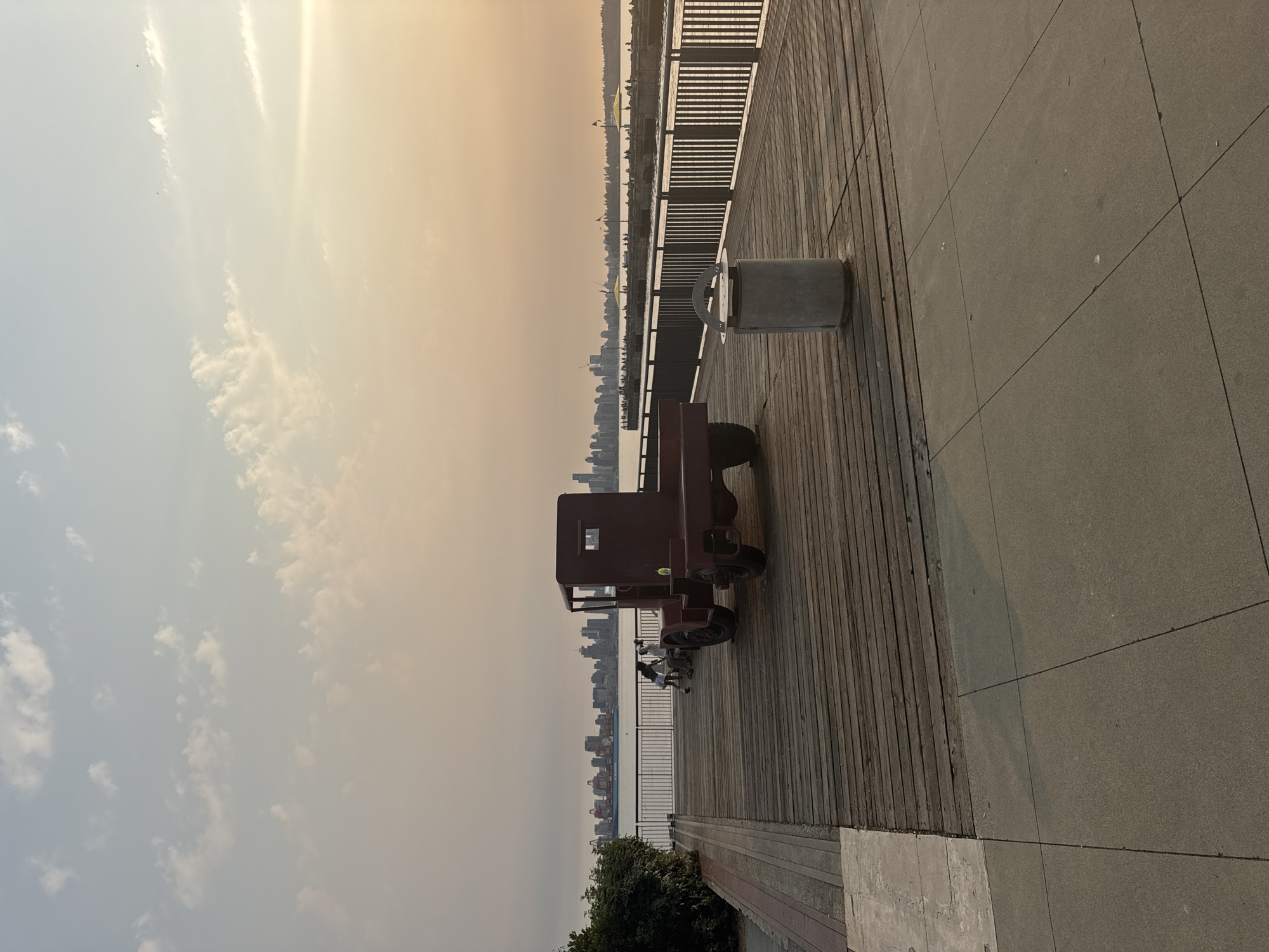 This image depicts a waterfront promenade in North Vancouver, Canada, likely during the late afternoon or early evening, given the soft, warm light. The foreground is dominated by a textured concrete pathway, transitioning to a wooden boardwalk. A large, vintage-style dark red truck chassis, devoid of a cab or wheels beyond its rear set, is prominently positioned on the boardwalk. To the right of the truck, a grey, cylindrical trash receptacle stands.
In the middle ground, the wooden boardwalk continues towards a metal railing that separates it from the water. Several people are visible on the boardwalk near the truck, some seated and others standing, engaged in conversation or leisure. Beyond the railing, the waters of the Burrard Inlet are visible, with a large cargo ship in the distance. Across the water, the skyline of Vancouver is clearly discernible, featuring numerous buildings and skyscrapers. The sky above is a gradient of pale blue and orange hues, with scattered white clouds. The overall atmosphere is calm and serene, suggesting a public space enjoying the day's end.