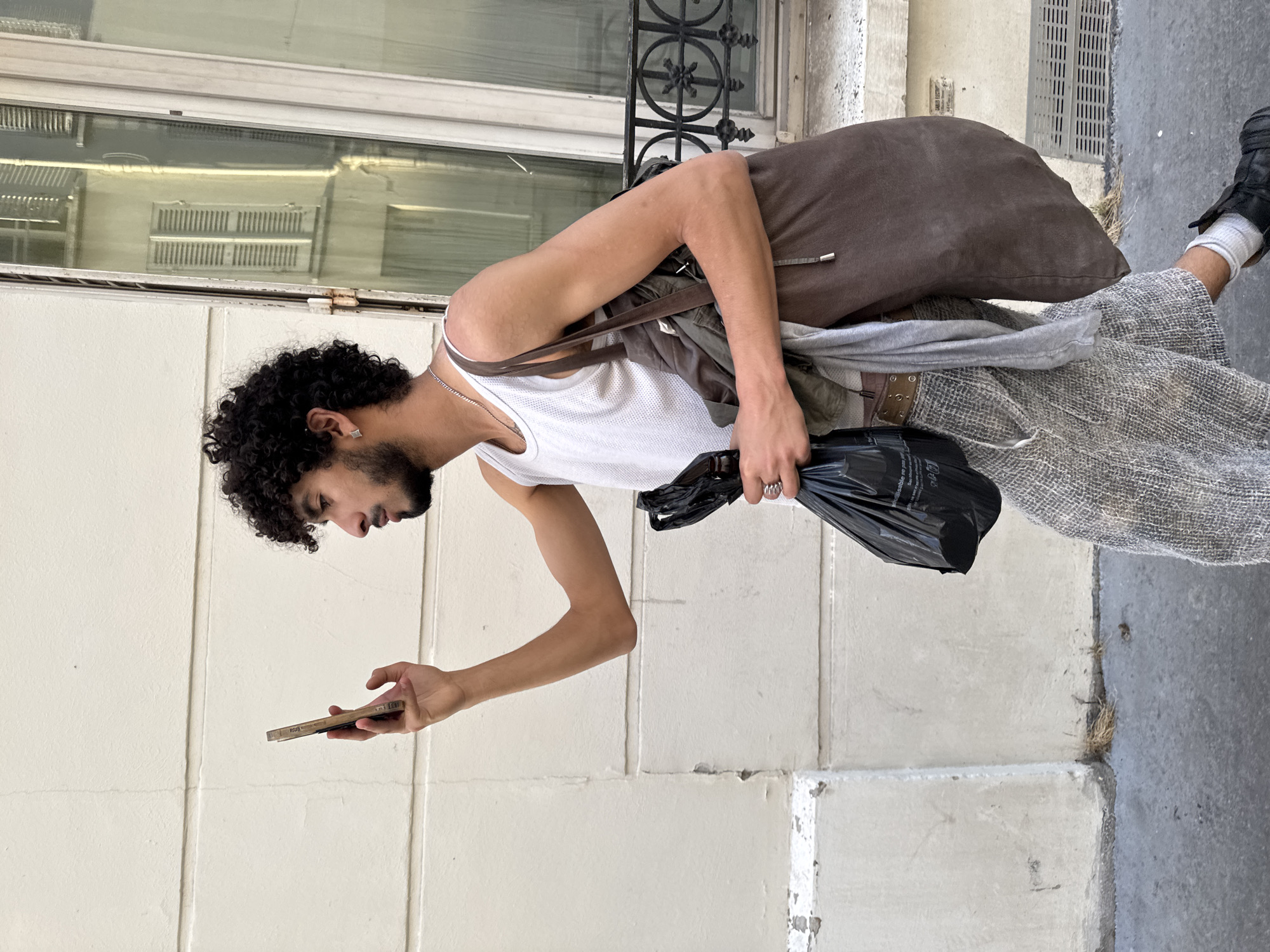 A young man with dark, curly hair, facial hair, and a silver chain is standing in front of a light-colored building, possibly in Paris, France, as suggested by the building's architecture. He is wearing a white tank top, wide, light-colored, patterned trousers, and black shoes with white socks. He carries a brown bag over his shoulder, another grey garment, and holds a black plastic bag in one hand while looking at a cellphone in the other. The background includes a window with a decorative iron grate and a sidewalk.
