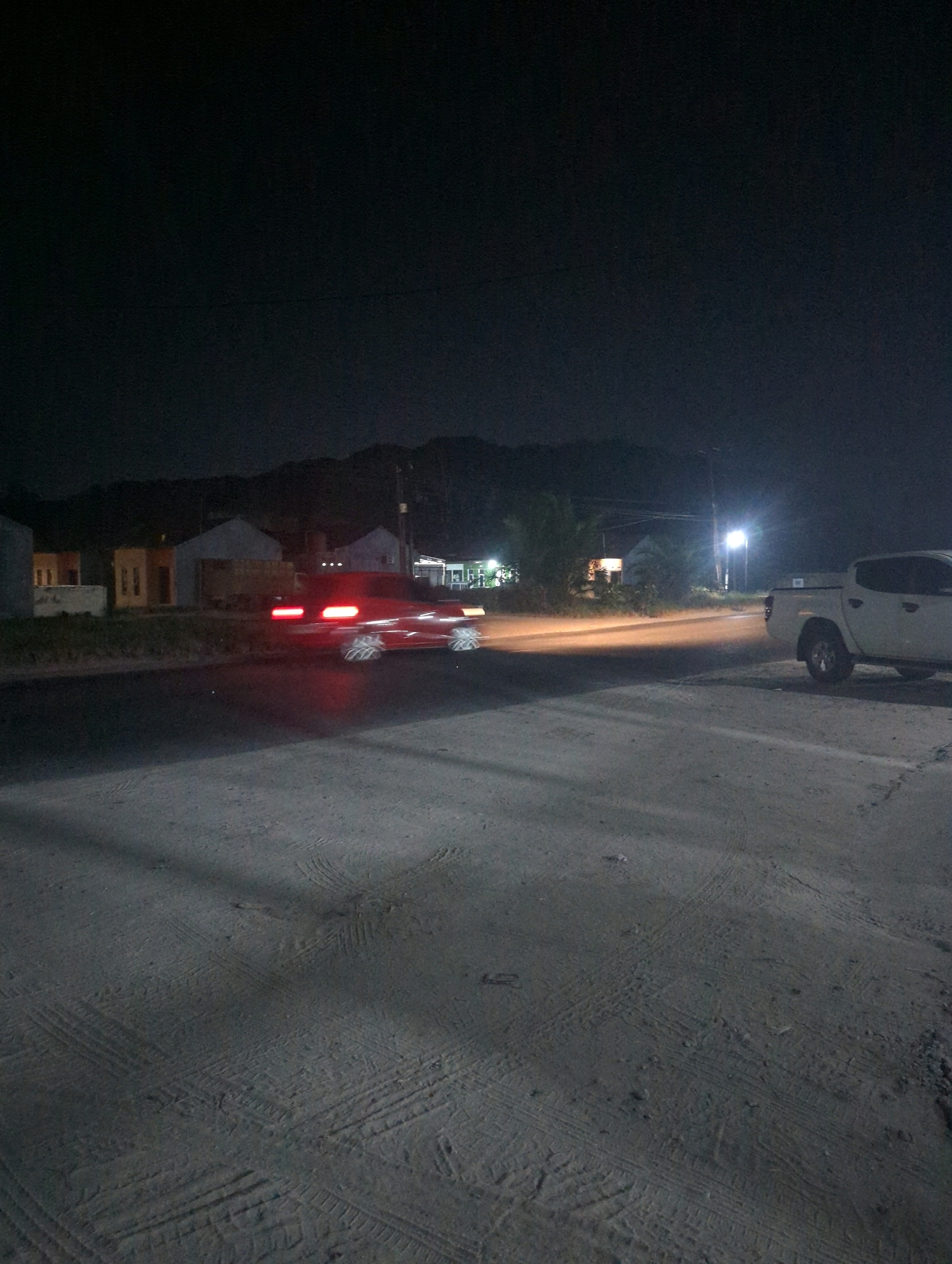 The image depicts a nighttime scene on a road in Batu Belaman, Indonesia. A red car is captured in motion, its taillights and wheels blurred due to a long exposure, indicating it is driving past. On the right side of the road, a white pickup truck is parked, its headlights off. The background shows a row of houses illuminated by dim lights, and further back, a hilly landscape under a dark sky. The road surface is paved and dusty, with visible tire tracks. A street light is visible in the distance, casting a glow on the road.