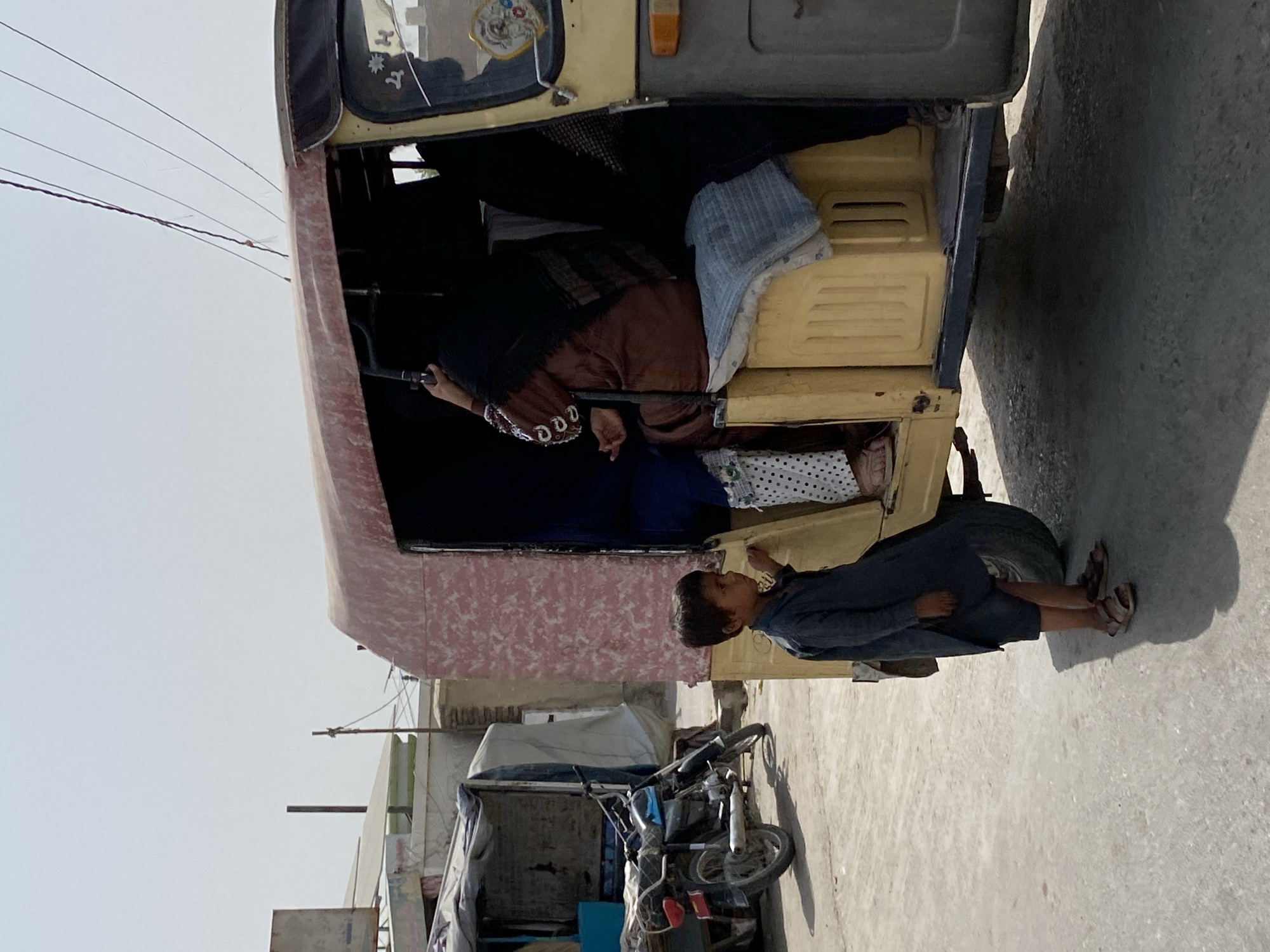 A young boy stands on a dusty street in Kandahar, Afghanistan, looking at a passenger auto-rickshaw. The boy, wearing a dark blue tunic and shorts with sandals, stands to the left of the vehicle. Inside the auto-rickshaw, several women are seated, some covered with black hijabs. One woman in a brown tunic is visible reaching for something. The auto-rickshaw has a faded pink and cream colored canopy and a yellow passenger compartment. To the left of the auto-rickshaw is a parked motorcycle. Buildings and utility poles are visible in the background under a clear, hazy sky.