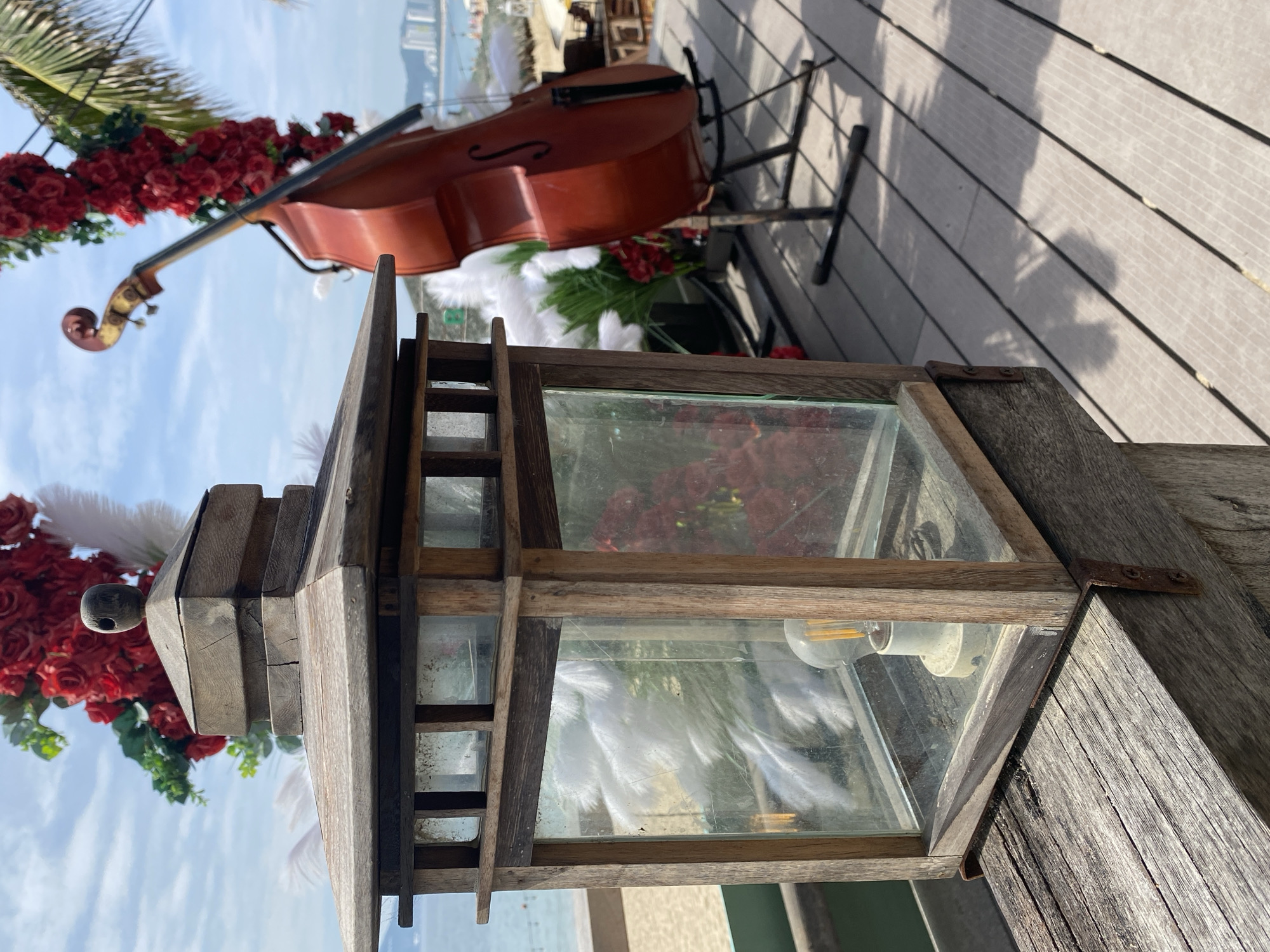 The image depicts a close-up of a wooden lantern on a weathered wooden deck. The lantern has glass panes, revealing a light bulb inside and some decorative white plumes. In the background, out of focus, is a cello, a floral arrangement of red roses, and a glimpse of the ocean with a beach and distant buildings, suggesting a coastal location. The scene appears to be set up for an event or performance, possibly at a resort or outdoor venue in Nha Trang, Vietnam, given the presence of palm trees and the distinct architectural style of the distant buildings. The lighting suggests it is daytime, with natural sunlight casting shadows. There are no people visible in the frame.