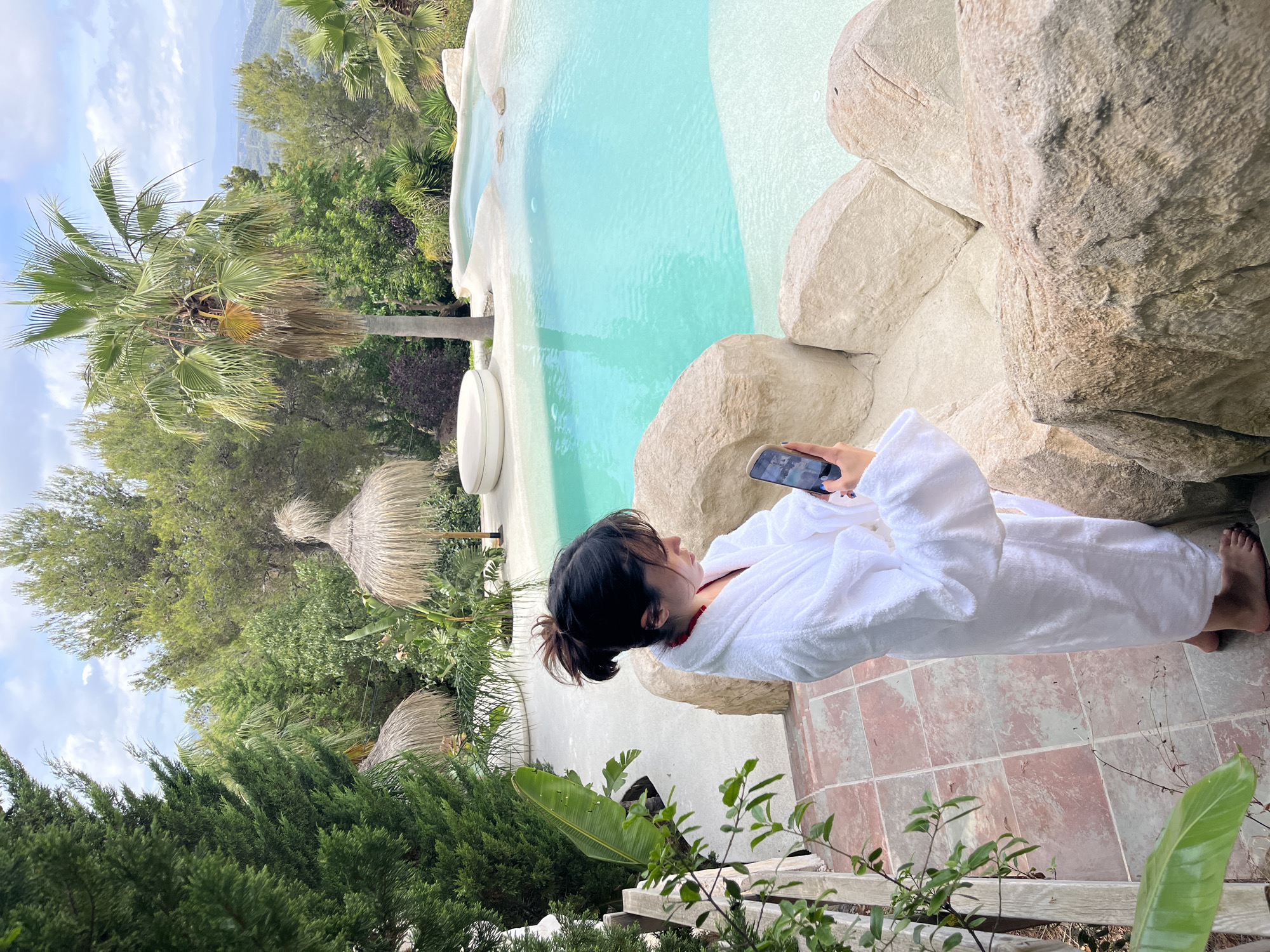 A young woman, seen from the shoulders down, stands on a tiled patio wearing a white bathrobe and holding a smartphone. She is positioned on the edge of an ornate, irregularly shaped swimming pool with turquoise water. The pool is surrounded by large, light-colored rocks and has a white, round, low-lying structure in the background. The scene is lush with abundant greenery, including palm trees, cypress trees, and tropical foliage. In the distance, rolling hills and mountains are visible under a cloudy sky. The location is specified as Torrelles de Llobregat, Spain.
