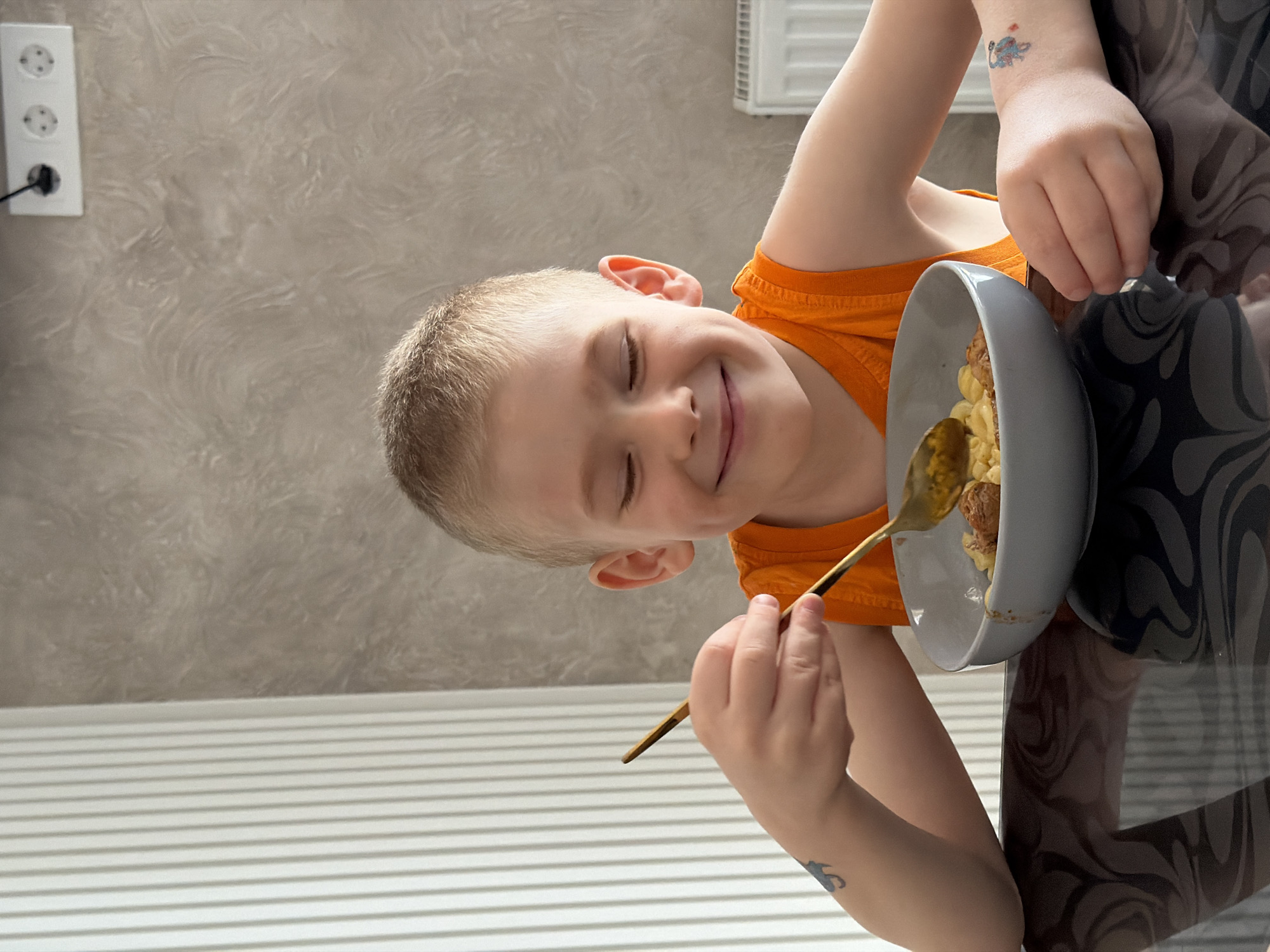 A fair-skinned child wearing an orange tank top sits at a reflective table with a gray bowl containing food and a golden spoon. The child is fair-skinned and has short, light blond hair. Their eyes are closed, and they are smiling. A tattoo is visible on the child's left wrist. The bowl of food appears to contain macaroni and meatballs. The table is black and reflective, showing a swirled pattern. A wall with a textured gray and white pattern is in the background, along with a white outlet and a white radiator. The location is Rossiyskiy, Russia.