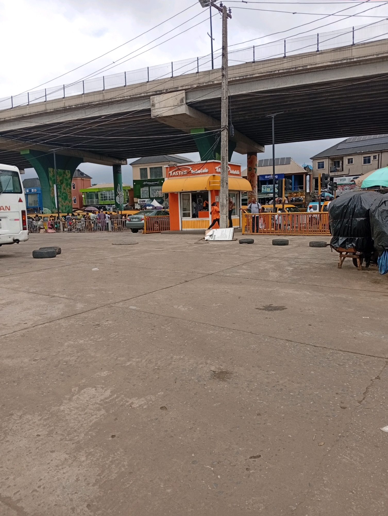 The image shows an outdoor scene in Lagos, Nigeria, under a large concrete overpass. In the foreground, the ground is paved with concrete, with several worn tires placed on it. To the right, a wooden cart covered with black plastic bags is visible.
In the middle ground, a bright orange and yellow kiosk with the sign "TASTES" is prominent. People are gathered behind a yellow barricade in front of the kiosk and other businesses. Some are waiting, while others appear to be interacting. A person in an orange shirt is walking away from the kiosk. A bus is partially visible on the far left of the image.
The background reveals various buildings, some with solar panels on their roofs, and streetlights. The overpass dominates the upper portion of the image, with visible infrastructure including power lines and a fence along its edge. The sky is overcast, indicating a cloudy day. The overall scene suggests a bustling urban environment, likely a transport hub or market area under an elevated road.