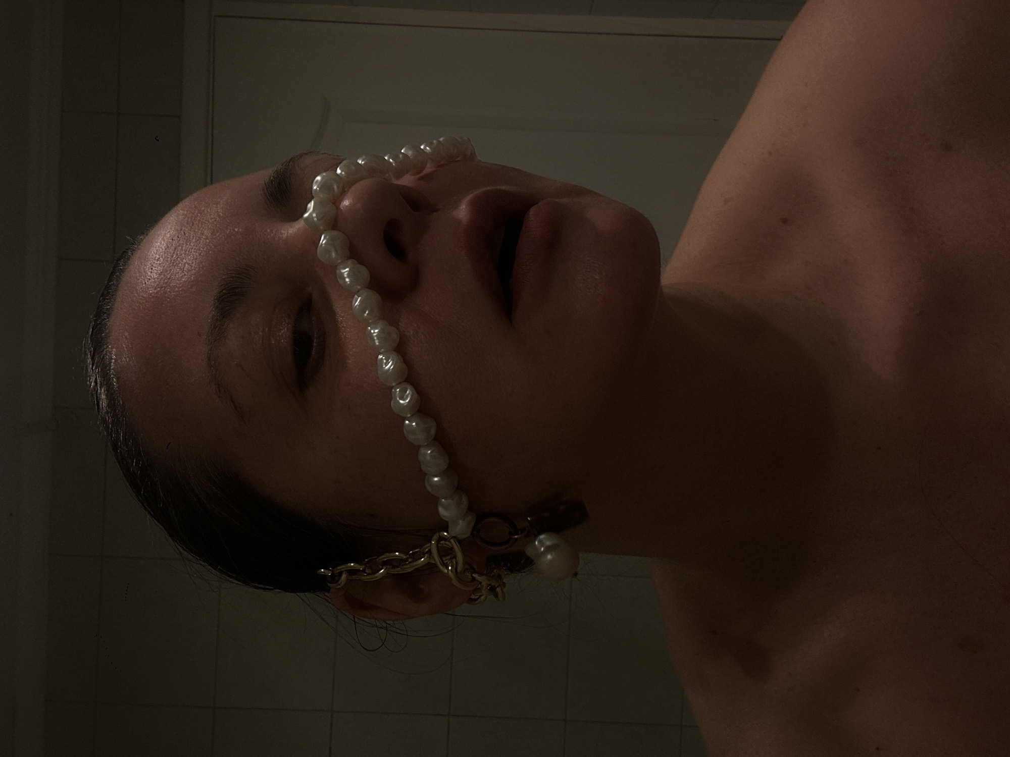 A close-up, low-angle shot shows a person's face and upper torso against a tiled background. The person has dark hair pulled back, exposing a forehead and skin with visible pores and freckles. The person is wearing a pearl necklace that drapes across their face from forehead to ear. Additionally, they have a gold chain and pearl earring on their left ear. The background consists of a white tiled wall and a door, suggesting a bathroom setting. The lighting is dim. The location is not evident from this image.