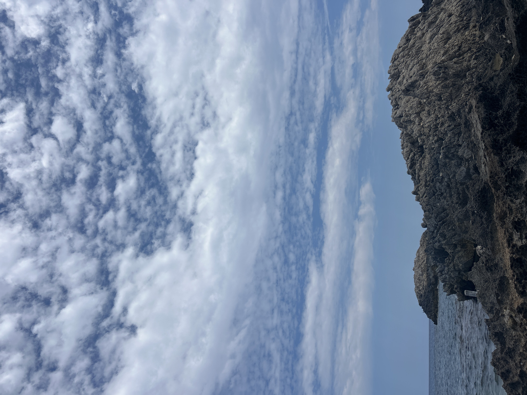 A wide-angle, eye-level shot captures the rugged coastline of Cala Lliteras, Spain, under a sky filled with scattered white clouds. The sea, a deep blue, gently laps against a dark, textured rocky shoreline that dominates the foreground and midground. In the distance, a larger rock formation juts out into the water, its surface weathered and uneven. The sky is a vibrant blue, with clouds forming an intricate pattern of white wisps and larger formations, creating depth and texture. The overall scene is one of natural beauty and tranquility.