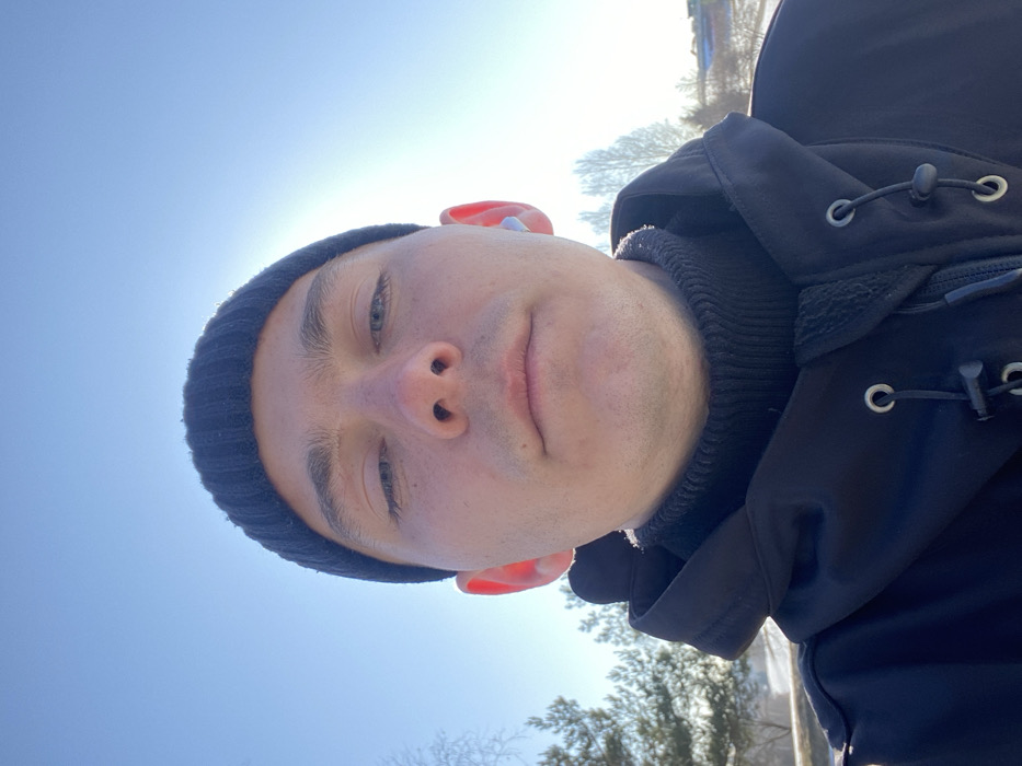 A close-up, low-angle selfie displays an adult male outdoors on a bright, sunny day. The subject, with light skin and a neutral expression, wears a black ribbed beanie, a black turtleneck sweater, and a black zippered jacket with visible drawstrings and metallic eyelets. A light-colored wireless earbud is present in his right ear. The background consists of a clear blue sky with a prominent sun flare in the upper right. Below the horizon, snow-covered ground is visible, flanked by bare deciduous trees and evergreen conifers. Faint structures, possibly houses or buildings with snow-covered roofs, are discernible in the distant background on the right, suggesting a semi-rural or park-like environment. The scene is consistent with a winter or early spring setting in a natural area. No specific landmarks are visible to precisely identify a location within the city of Moscow, Russia.