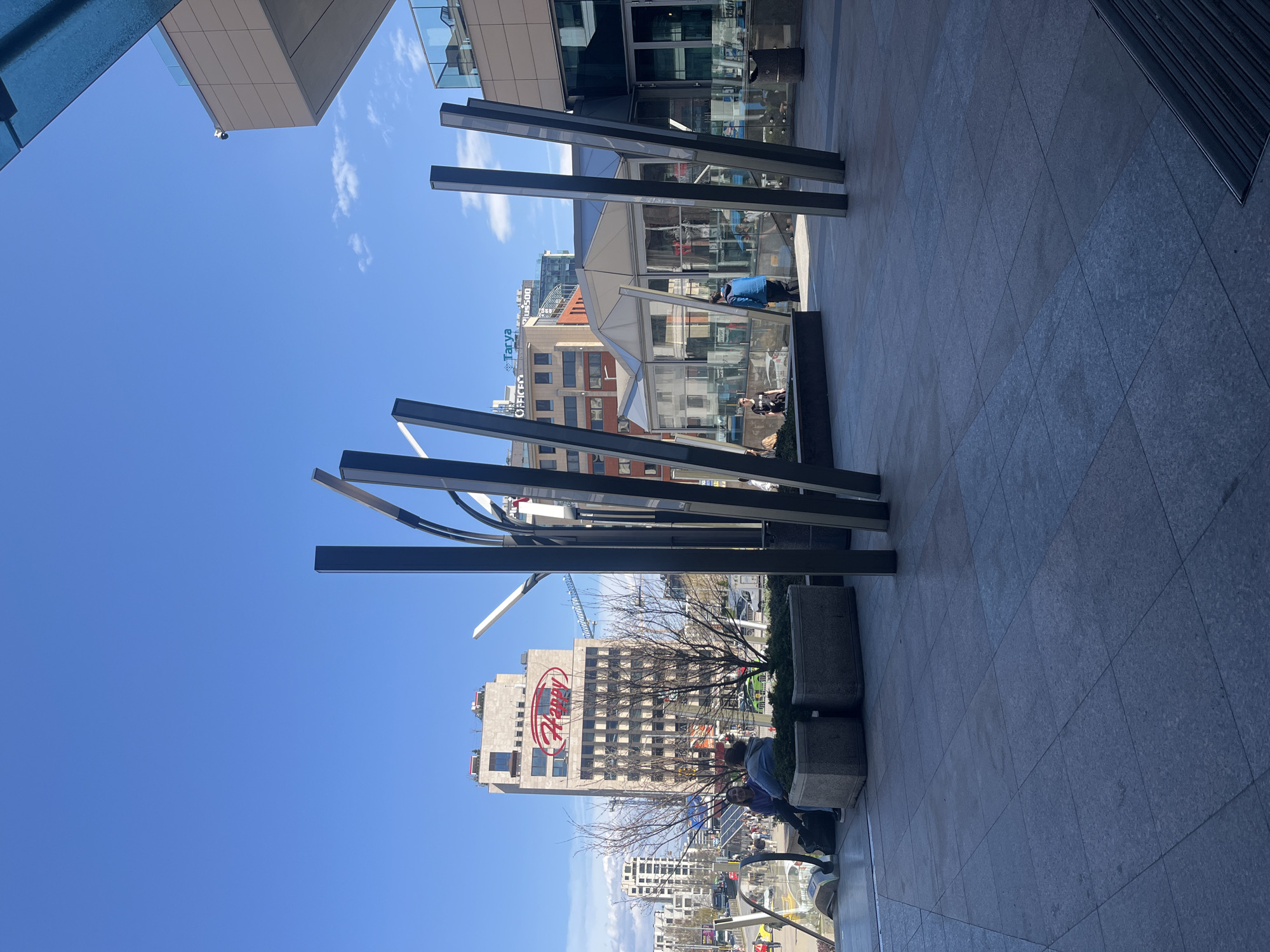The image depicts an outdoor urban scene in Sofia, Bulgaria, under a clear blue sky. Foregrounded are modern architectural features consisting of tall, dark, metal posts, some connected by curved, reflective elements, seemingly part of a decorative installation. The ground is paved with large, square tiles, leading into a pedestrian area with several people visible. Buildings are visible in the background, including a high-rise bearing a large "Hyundai" logo. There are also smaller glass structures, possible bus shelters or display cases, where individuals can be seen. Plant boxes with some greenery adorn the area. There is a crane near the tall building. The composition suggests a perspective from a low angle, looking upwards towards the sky and the buildings.