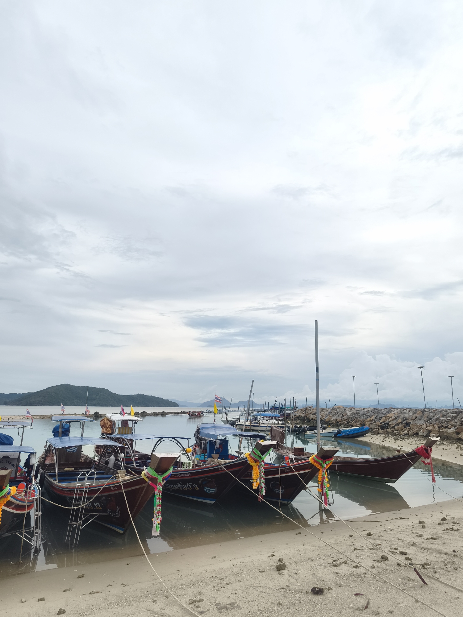 The scene depicts a group of traditional Thai longtail boats moored at the beach in Baan Thong Krut, Thailand. The boats, which are predominantly brown and maroon with blue canopies, are adorned with colorful floral garlands at their prows. They are tied together with ropes that extend onto the sandy beach. The water in the harbor is calm, reflecting the overcast sky. A rock breakwater stretches into the water, with several light poles visible on it. In the background, a hilly coastline can be seen under the cloudy sky.