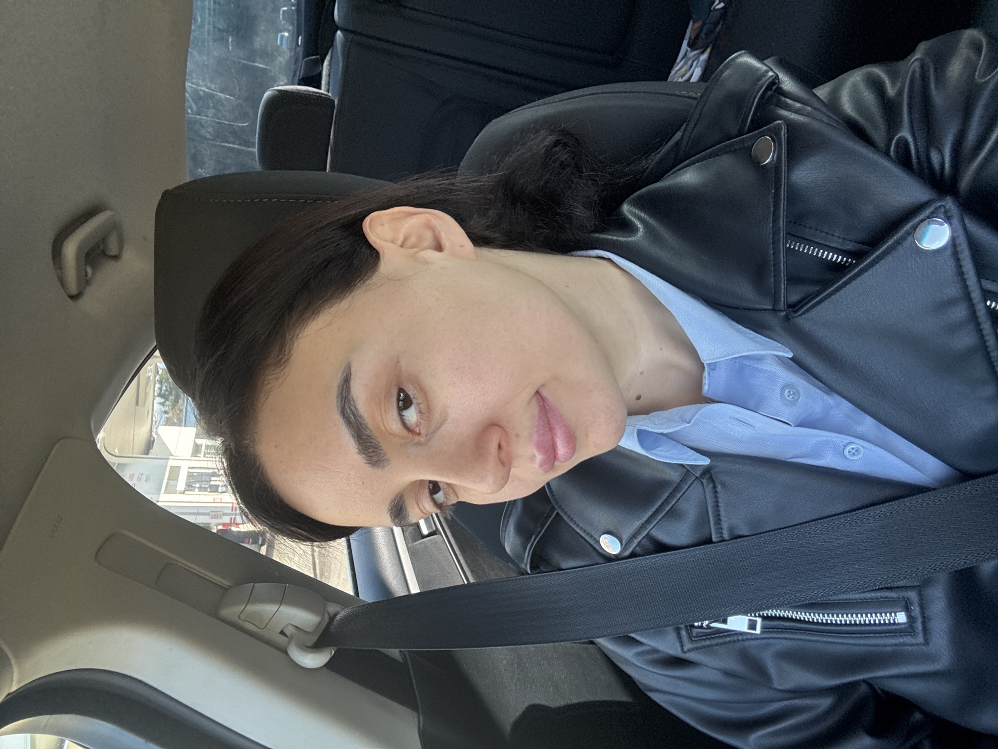 A woman is seated in a car, wearing a black leather jacket over a light blue button-down shirt, with her seatbelt fastened. The surrounding environment suggests a city location. The window reveals urban structures, possibly in Tbilisi, Georgia, though specific landmarks are not visible. The interior of the car, including the seat, headrest, and dashboard elements, are discernible. The woman appears to be looking towards the camera.