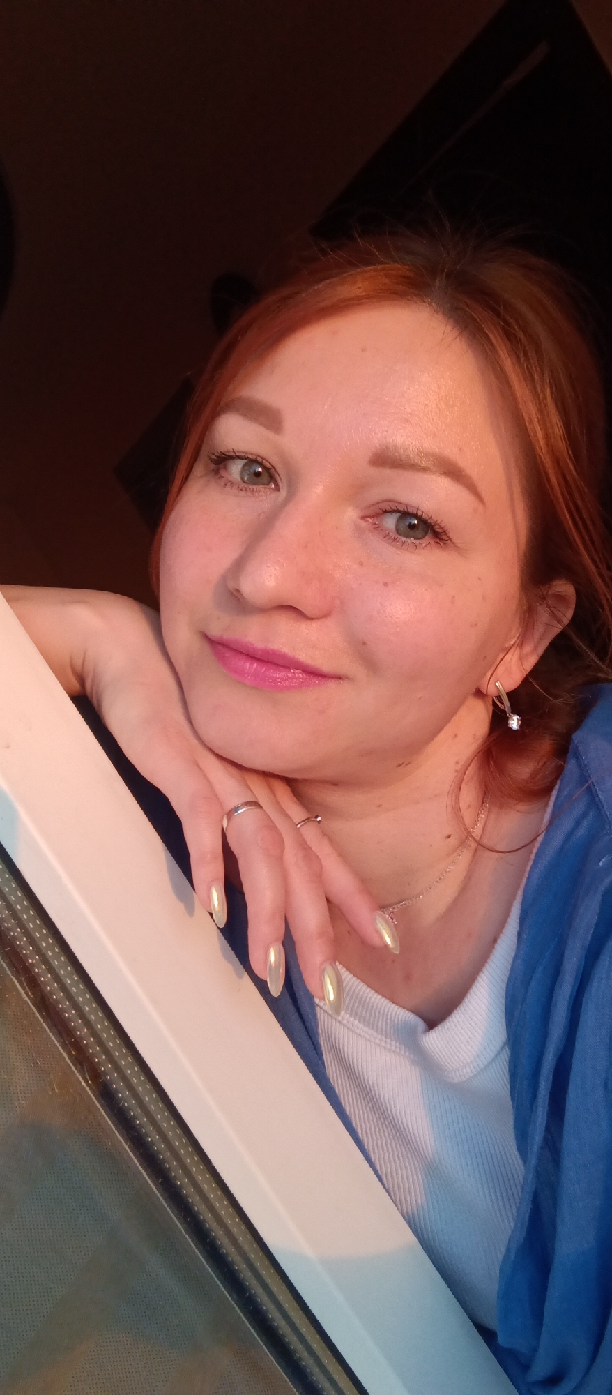 A close-up shot of a woman with reddish hair and fair skin is looking at the camera with a slight smile. She is wearing pink lipstick, a white ribbed top, and a blue garment draped over her shoulder. She has jewelry including earrings, rings, and a delicate necklace. Her nails are long and painted with an iridescent color. The background is dark, except for a window frame where her hand rests. The image location is in the city of Unknown, Unknown.