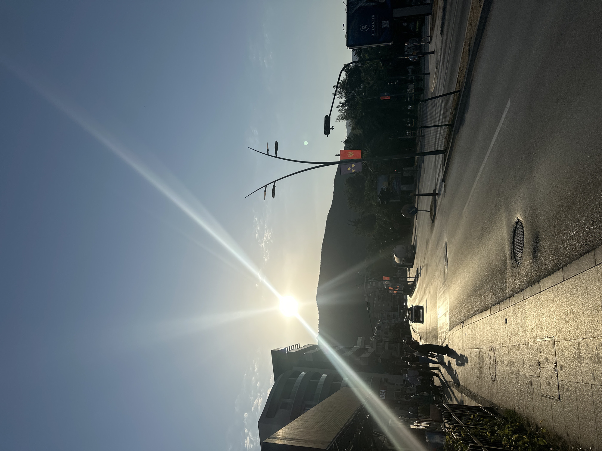 The image captures a sun-drenched street scene in Budva, Montenegro, during what appears to be late afternoon or early evening, judging by the low angle of the sun and the long shadows cast. The sun is positioned high and bright in the sky, creating strong lens flares and a golden hue across the pavement and buildings. On the left, a modern, multi-story building with a dark facade and balconies dominates the foreground. Its lower level features shops, with a sign for "IDEA" visible. A sidewalk runs alongside the building, populated by several individuals, some walking and others standing. A metal fence borders the sidewalk. The street itself is paved and appears to be a main thoroughfare, with a few vehicles visible, including a car in motion and a truck further down the road. Streetlights and traffic signals line the road, and two prominent street lamps with multiple lights stand out. Red flags with yellow emblems are displayed on poles along the street. In the background, a dark, tree-covered hill rises, silhouetted against the bright sky. The overall atmosphere is one of urban activity bathed in the warm, intense light of the setting sun.