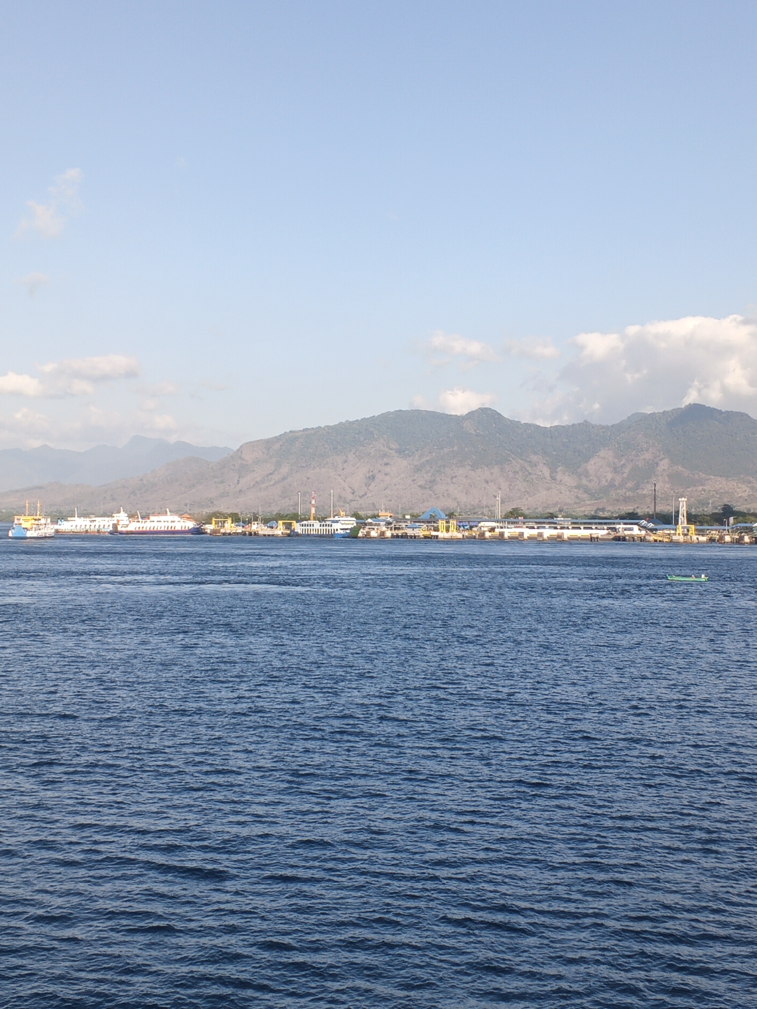 The image depicts a harbor scene in Melaya, Indonesia, under a clear blue sky with scattered white clouds. The foreground is dominated by the deep blue, rippling water of the sea. In the mid-ground, a busy harbor stretches across the frame, featuring several large ferries docked and possibly in transit. These ferries are white with colorful accents, and some have multiple decks. Adjacent to the ferries, there is a port infrastructure with buildings, piers, and cranes. The background is characterized by a range of mountains, their slopes appearing dry and covered with sparse vegetation. A small, green boat with two figures aboard is visible in the water on the right side of the image, adding a sense of scale and activity. The overall impression is one of a functional port city set against a dramatic natural backdrop.