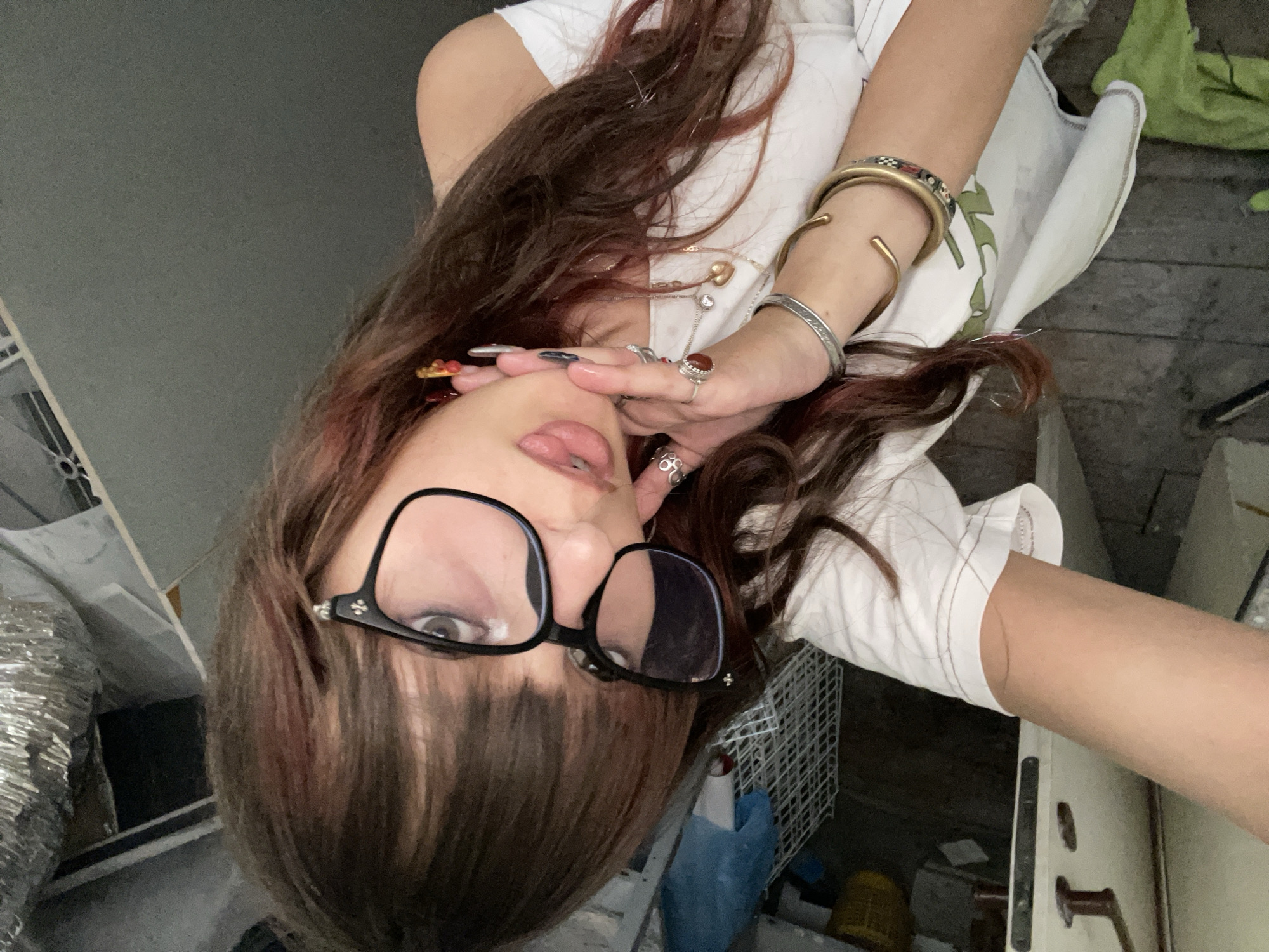 The image shows a young woman with long, dark hair with reddish highlights, wearing black-rimmed glasses and a white off-the-shoulder top. She is holding her right hand up to her face, with several rings on her fingers. She is also wearing several bracelets on her left wrist. Her tongue is sticking out slightly. The background appears to be an indoor space with industrial-looking elements, such as exposed pipes and shelving. There are also some plastic containers and what looks like a laundry basket. The floor is made of rough wooden planks. The image is taken from a high angle, looking down at the woman. The lighting is somewhat dim, suggesting an indoor setting. No specific location in Raanana, Israel can be identified from the visual cues in this image.
