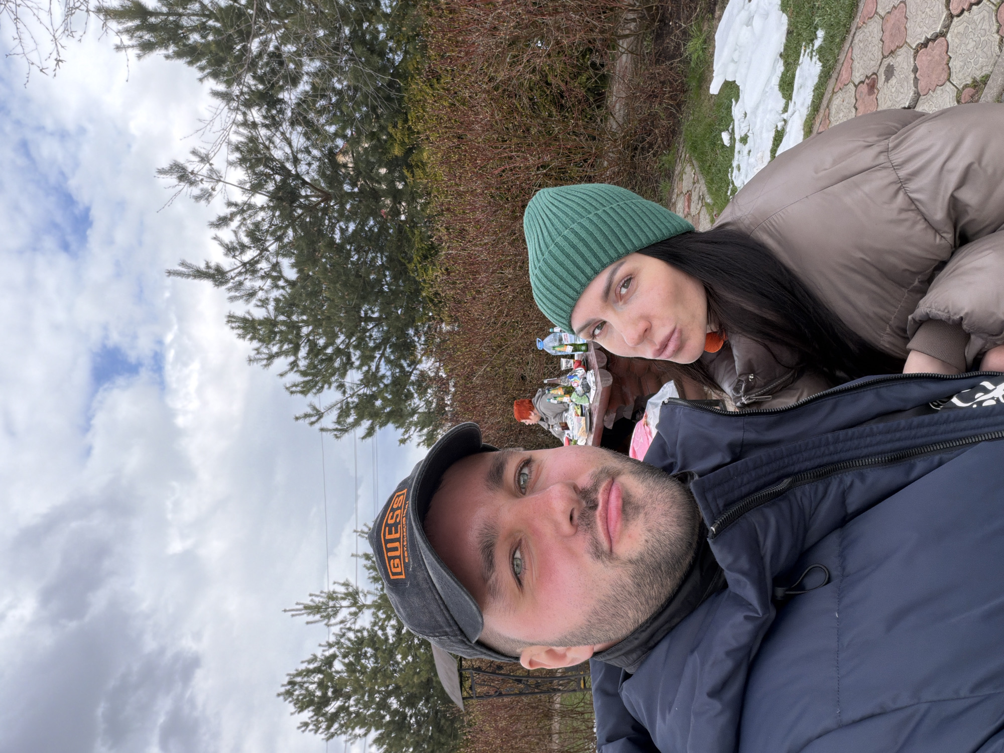 A selfie-style photograph captures a man and a woman outdoors, likely in late winter or early spring, evidenced by the patches of melting snow on the ground and their warm attire. The man, in the foreground on the left, is wearing a dark blue winter jacket and a dark baseball cap with the brand name "GUESS" visible in orange text. He has a beard, mustache, and light-colored eyes, looking directly at the camera with a neutral expression.
Behind him, on the right, the woman wears a brown puffer jacket and a green ribbed knitted beanie, with long dark hair visible underneath. A distinct orange earring or part of an earring is visible. She also looks towards the camera with a slightly serious or pouty expression.
In the background, positioned between the two subjects, a table is visible, laden with various items such as bottles, plastic bags, and other gathering remnants. A third person, possibly a child with red hair, is partially obscured at this table. The ground features a mix of light-colored pavers, grass, and remnants of snow. The surrounding environment consists of evergreen trees and bare deciduous bushes, suggesting a park or backyard setting. The sky above is overcast with prominent white and grey clouds, indicating a cloudy daytime. The scene is set in what appears to be a casual outdoor gathering in Lytkino, Russia.