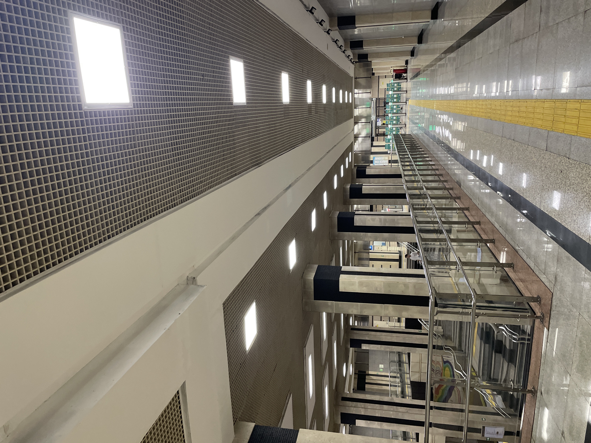 The image depicts an interior view of a modern metro station in Almaty, Kazakhstan. The scene is characterized by a long, wide corridor with a polished floor that reflects the numerous square and rectangular ceiling lights. The ceiling itself is a grid structure with integrated lighting panels. Massive concrete columns, accented with black vertical stripes, support the elevated structure.
On the left side of the corridor, a staircase with a glass and stainless steel railing descends. The stairs have a patterned design and a mural is visible on the wall beneath the staircase. On the right side of the corridor, a prominent yellow tactile paving strip runs along the edge of the walkway, indicating a pathway for visually impaired individuals.
In the distance, at the end of the corridor, a row of turnstiles with green tops suggests an exit or entrance area. A few people are visible in the background, including one individual standing near the turnstiles, and another person walking down the stairs on the left. The overall environment is clean, well-lit, and designed with a contemporary aesthetic, typical of public transportation hubs. The architecture suggests a functional and orderly space.