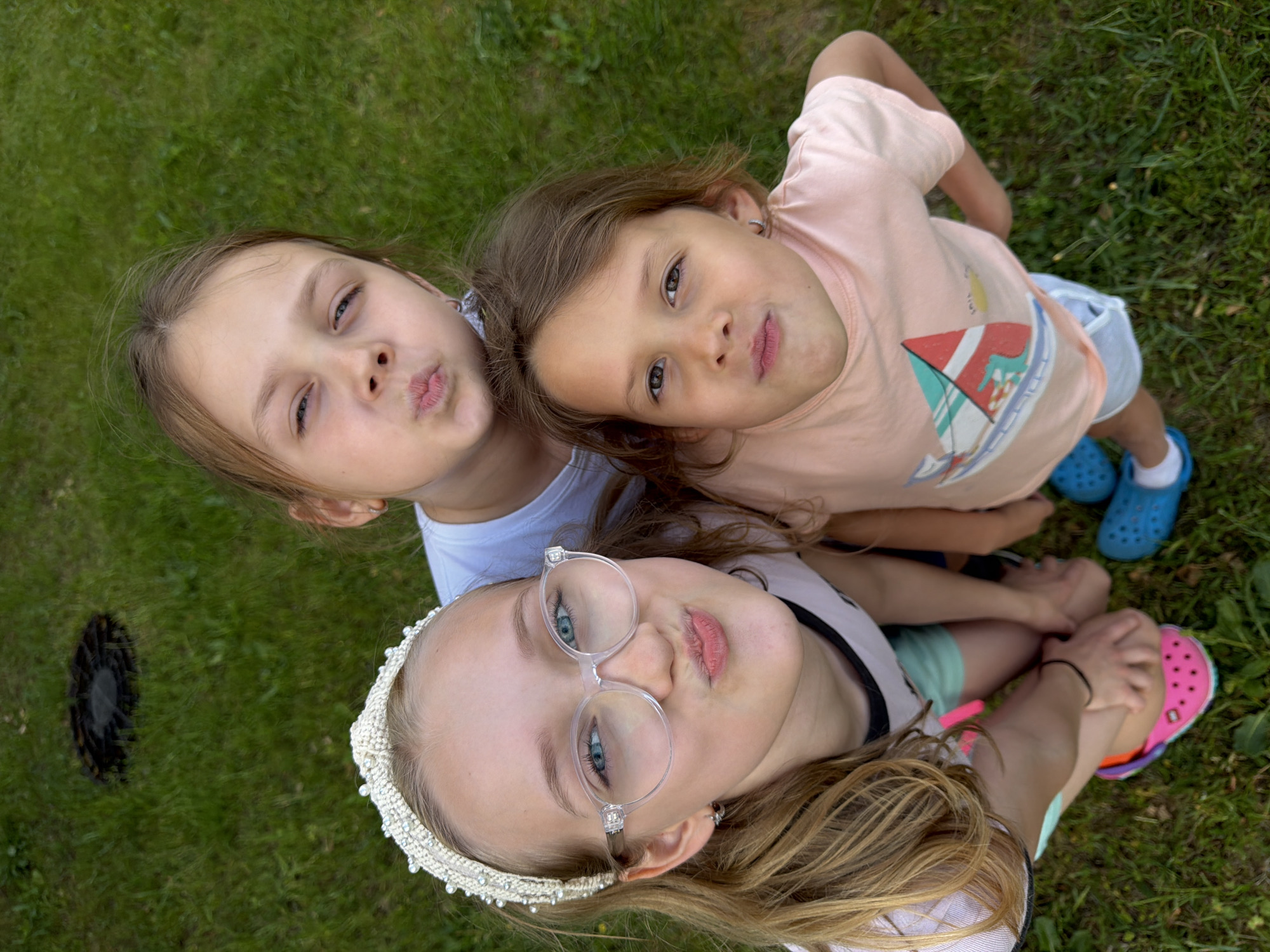 The image displays three young females outdoors in Dreiliņi, Latvia, captured from a high-angle perspective with all subjects looking upward towards the camera. In the foreground, positioned left of center, a girl with blonde hair, wearing a white beaded headband and clear-framed eyeglasses, makes a puckering expression with her lips. She is dressed in a white short-sleeved top with black trim, light green shorts, and pink Crocs. Directly behind her and slightly to the right, a second girl with light brown hair also presents a puckering expression and wears a white short-sleeved top. To the right of the central girl, a third, younger female with light brown hair wears a peach-colored t-shirt with a colorful sailboat graphic and blue Crocs; her facial expression is neutral with slightly pursed lips. The ground is uniformly covered in green grass. In the upper background, a dark, circular utility cover is visible embedded within the grass.