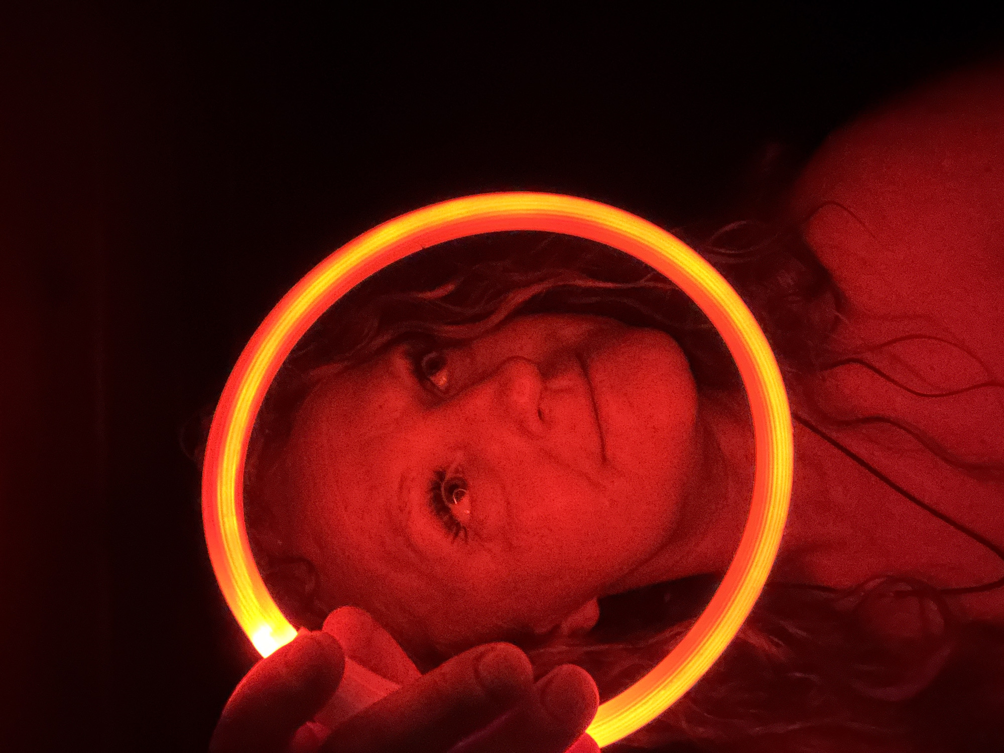 This close-up portrait features a person in a dimly lit or dark environment, likely at night or indoors. A bright, circular glow stick or LED light ring, radiating a vibrant yellow and orange light, frames the person's face. The intense light source bathes the subject's skin and immediate surroundings in a strong reddish-orange hue, creating a dramatic and warm cast across the scene.
The person's head is tilted, and they gaze directly into the camera with a neutral or slightly contemplative expression. Their eye is distinctly visible within the luminous circle, reflecting the light. Long hair is partially visible around their head and shoulders, blending into the rich red tones and dark background. Fingers are seen in the lower portion of the image, holding the glowing ring, positioning it carefully around the face. The composition emphasizes the interplay of light and shadow, highlighting the facial features within the glowing frame. The setting suggests an artistic exploration of light, color, and portraiture. This scene is set in Chaikovsky, Russia.