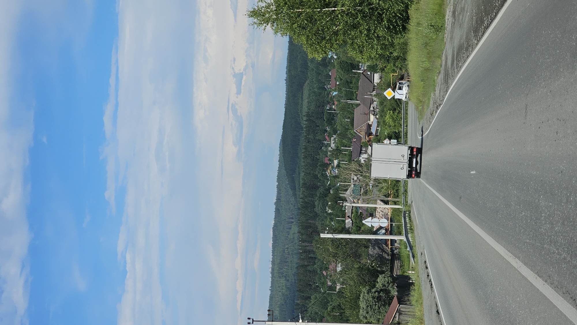 A white truck is driving on a paved road. The road curves to the right. On the left side of the road, there is a village with houses nestled amongst green trees and a dense forest. The sky is blue with some white clouds. The sun is shining, suggesting it is daytime. There are several road signs visible, including a yellow diamond-shaped sign indicating a hazard or warning. The overall scene depicts a rural or semi-rural setting in Revda, Russia.