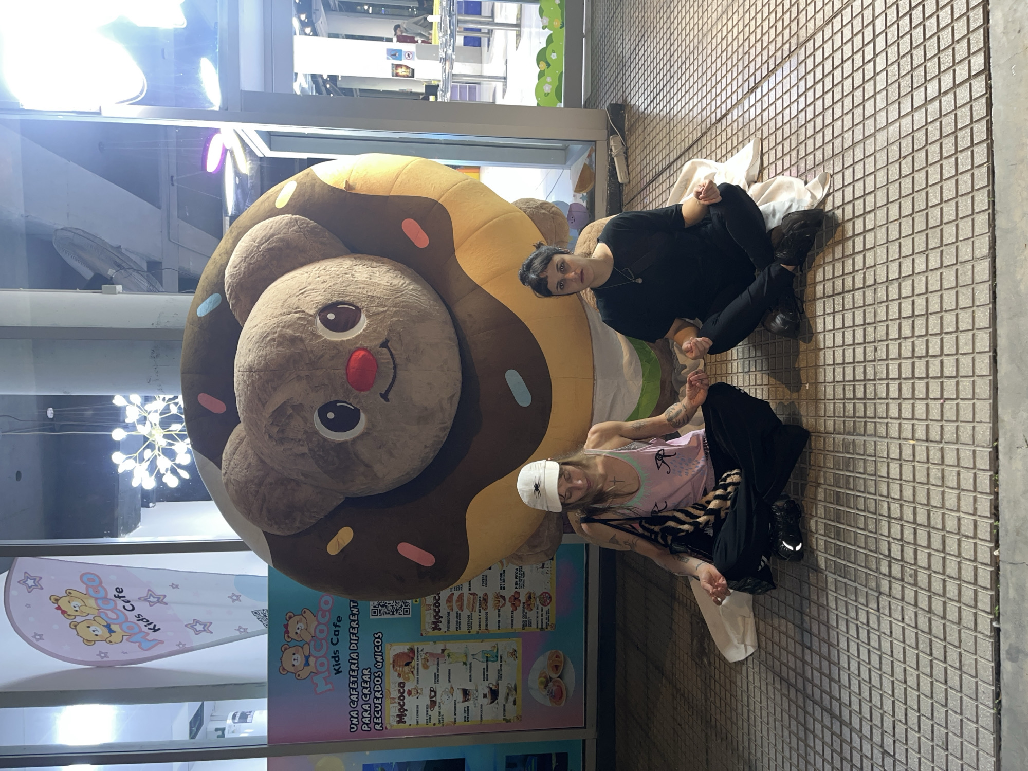 Two people are sitting on a tiled sidewalk next to a large plush toy shaped like a donut with a bear's face, likely in Buenos Aires, Argentina. The person closer to the camera is wearing a light pink tank top, black pants, and a white beanie, and has multiple tattoos visible on their arms. The other person is wearing black clothes. They are both positioned in front of a business advertising "Mococo Kids Cafe," suggesting they are near or in front of this cafe. The advertisement mentions it is a cafeteria that creates unique memories. The cafe's menu can be seen written on the windows. The background shows a glimpse of the interior of the building, with a hanging light fixture visible on the left. The sidewalk is made of square tiles.