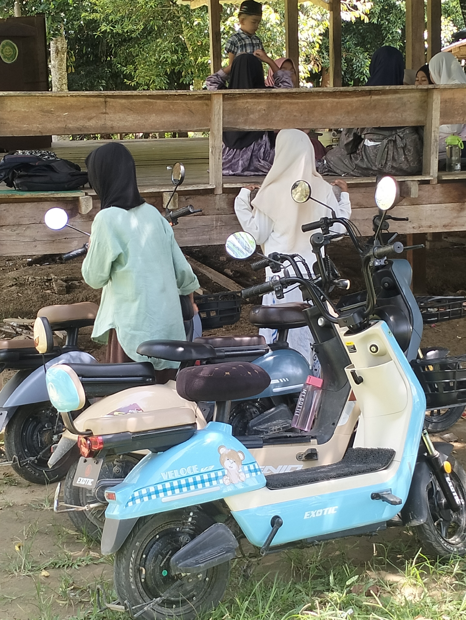 The image depicts an outdoor scene featuring several electric scooters parked on uneven ground with sparse grass and dirt. In the foreground, a light blue and cream-colored scooter prominently displays the brand "EXOTIC" and a cartoon bear graphic with checkered patterns. To its left, a beige and grey scooter is visible, followed by a darker grey scooter further back.
In the middle ground, two individuals are partially visible. One person, wearing a light green long-sleeved top and a black headscarf, stands beside a scooter. Another person, wearing a white hijab and long sleeves, is also present.
The background reveals a wooden structure, possibly a pavilion or an elevated platform, with several other people, including children, seated or standing. The environment is characterized by lush green trees suggesting a tropical or subtropical setting.
The scene appears to be a public gathering or a resting area, likely within a community in Meunasahalue, Indonesia, given the context of electric scooters being a common mode of transportation in such areas. The overall impression is one of casual activity and communal presence.