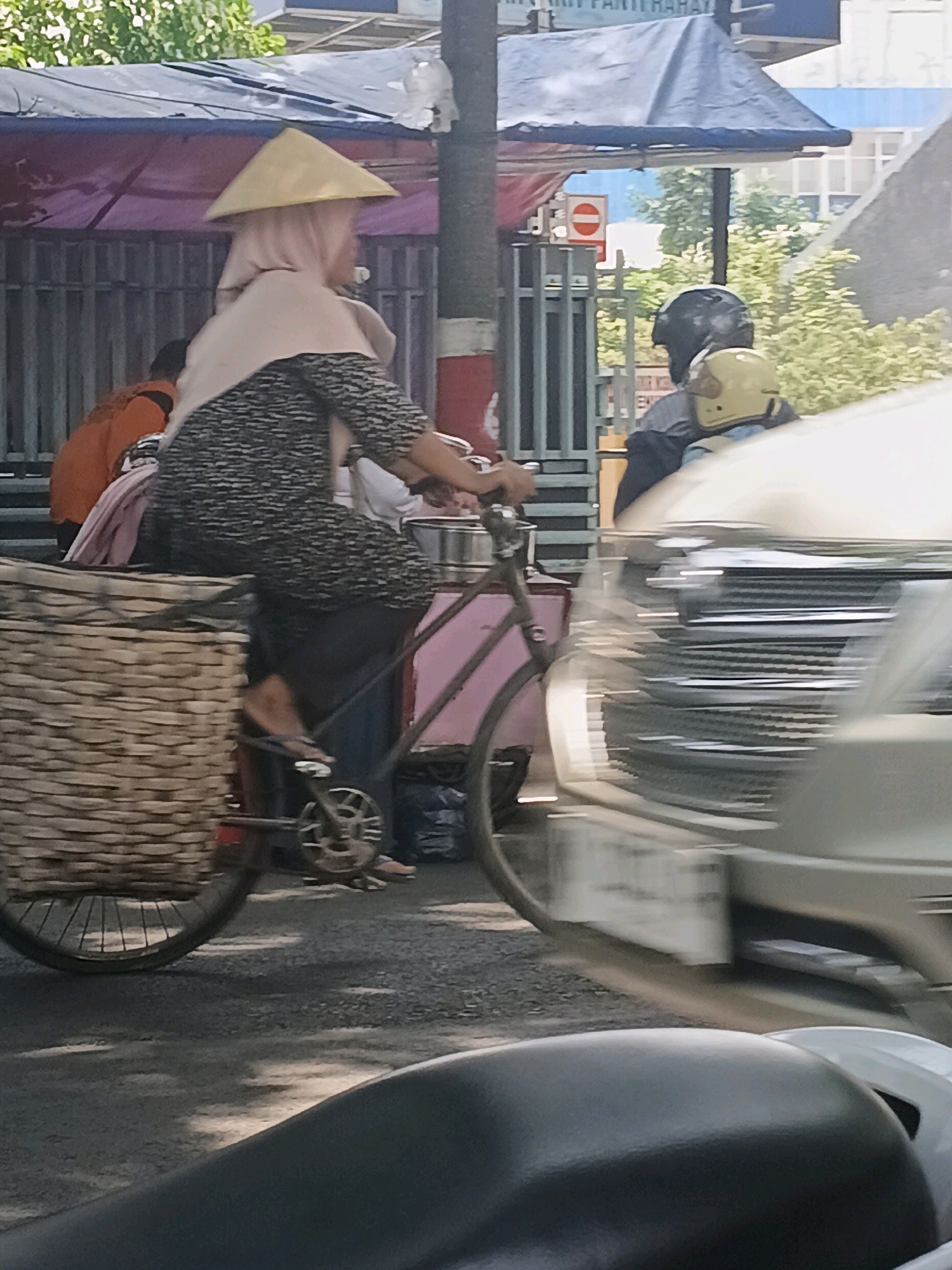 In Purwodadi, Indonesia, a woman wearing a conical hat and a hijab rides a bicycle. The bicycle has a large woven basket attached to the rear. The woman is dressed in a patterned long-sleeved shirt and dark pants. She is in motion on a paved road, with a vehicle in the foreground, partially blurred due to motion. In the background, there are some structures covered with tarps, metal fencing, trees, and other people, one wearing an orange shirt and another on a motorbike wearing a helmet. The scene appears to be a street or market area.