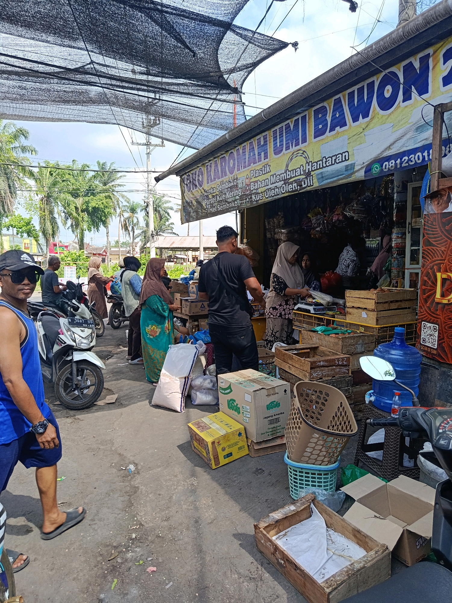 The image depicts a street-level view of a small shop in Jatiroto, Indonesia. A prominent banner above the shop reads "SANKO KAROMAH UMI BAWON 2", with additional text indicating "Aneka Sembako", "Plastik", "Bahan Membuat Hantaran", "Parsel Kue Kering", and "Kue Oleh Oleh". A phone number, "08123011", is also visible on the banner.
Several people are present at the scene. In the foreground, a man wearing a blue tank top, dark sunglasses, and a black baseball cap stands to the left, facing the camera. He is wearing dark shorts and grey flip-flops. To his right, a white motorcycle is parked.
Further into the scene, several individuals are interacting around the shop. A woman in a brown hijab and a patterned dress is standing near the shop entrance, appearing to be serving a customer. Another woman in a green hijab and a green patterned dress is standing to her left. Several other individuals, some on motorcycles and some on foot, are also visible.
The shop appears to be selling a variety of goods, including food items (sembako), plastics, and party supplies (hantaran). Several boxes, crates, and bags of goods are stacked outside the shop, suggesting it is a busy marketplace. Notably, a large blue water dispenser is placed to the right of the shop, along with a plastic basket and a motorcycle.
The environment is characteristic of a tropical climate, with palm trees visible in the background and a bright, overcast sky. The ground is paved with asphalt, and there are some debris scattered on the ground. A black mesh canopy is stretched over the area, providing shade. The overall impression is of a bustling local market scene in a residential or commercial area of Jatiroto.