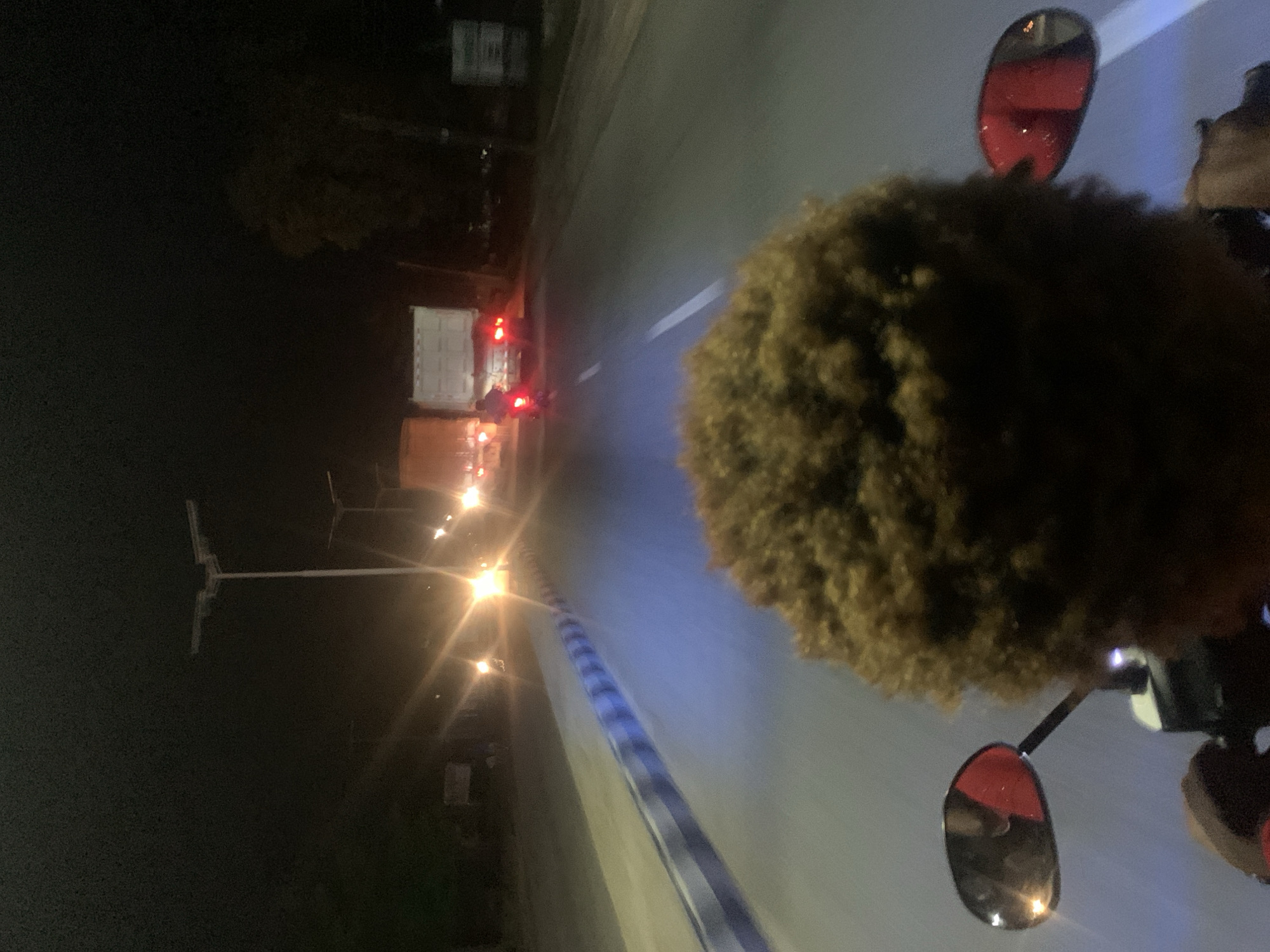 The image shows a person with an afro riding a motorcycle at night in Adankolo, Nigeria. The rider is facing away from the camera. The motorcycle has red mirrors. The road is illuminated by streetlights and headlights from a truck ahead. The environment is dark, with buildings and trees visible in the background.