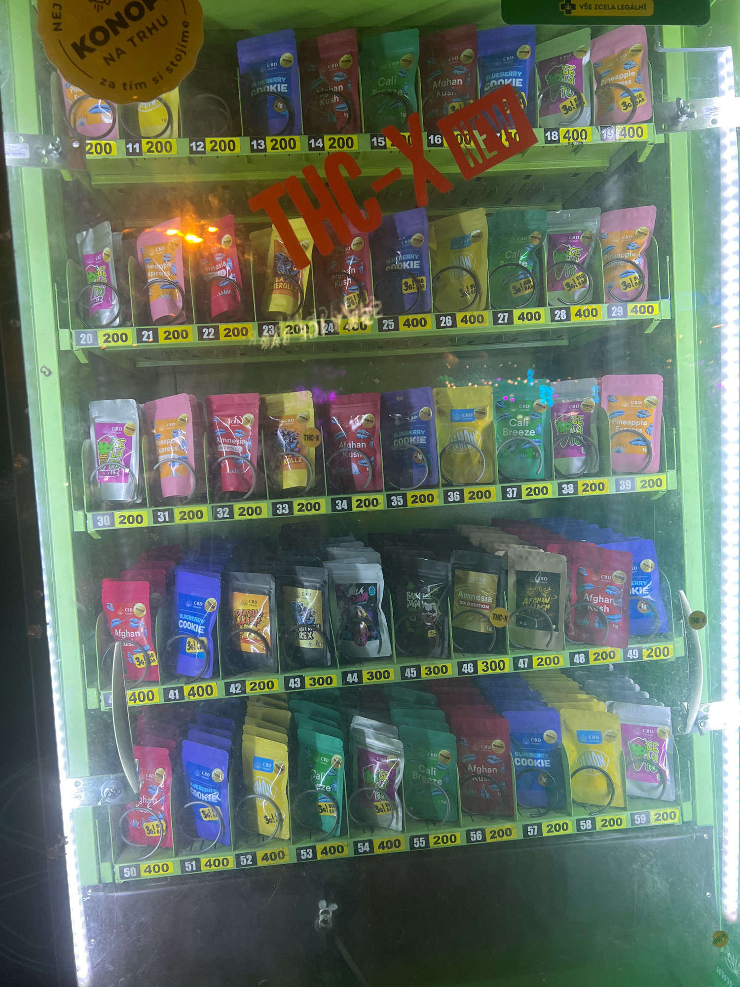 The image shows the interior of a green vending machine filled with various pouches of what appears to be cannabis products. The pouches are arranged in rows and columns, each with a product name and a price displayed below it. Some of the product names visible include "Blueberry Cookie," "Afghan Kush," "Cali Breeze," and "Pineapple Express." The vending machine is located in Brno, Czechia, as indicated by the text "KONOPÍ NA TRHU za tím si stojíme" which translates to "Cannabis on the market, we stand behind it." There is a red "NEW" sticker on some of the products. The lighting in the image is bright, illuminating the products clearly. The background is dark, suggesting it might be nighttime or an indoor setting. There are no people visible in the image. The overall scene depicts a commercial vending machine offering cannabis-related items in Brno.
