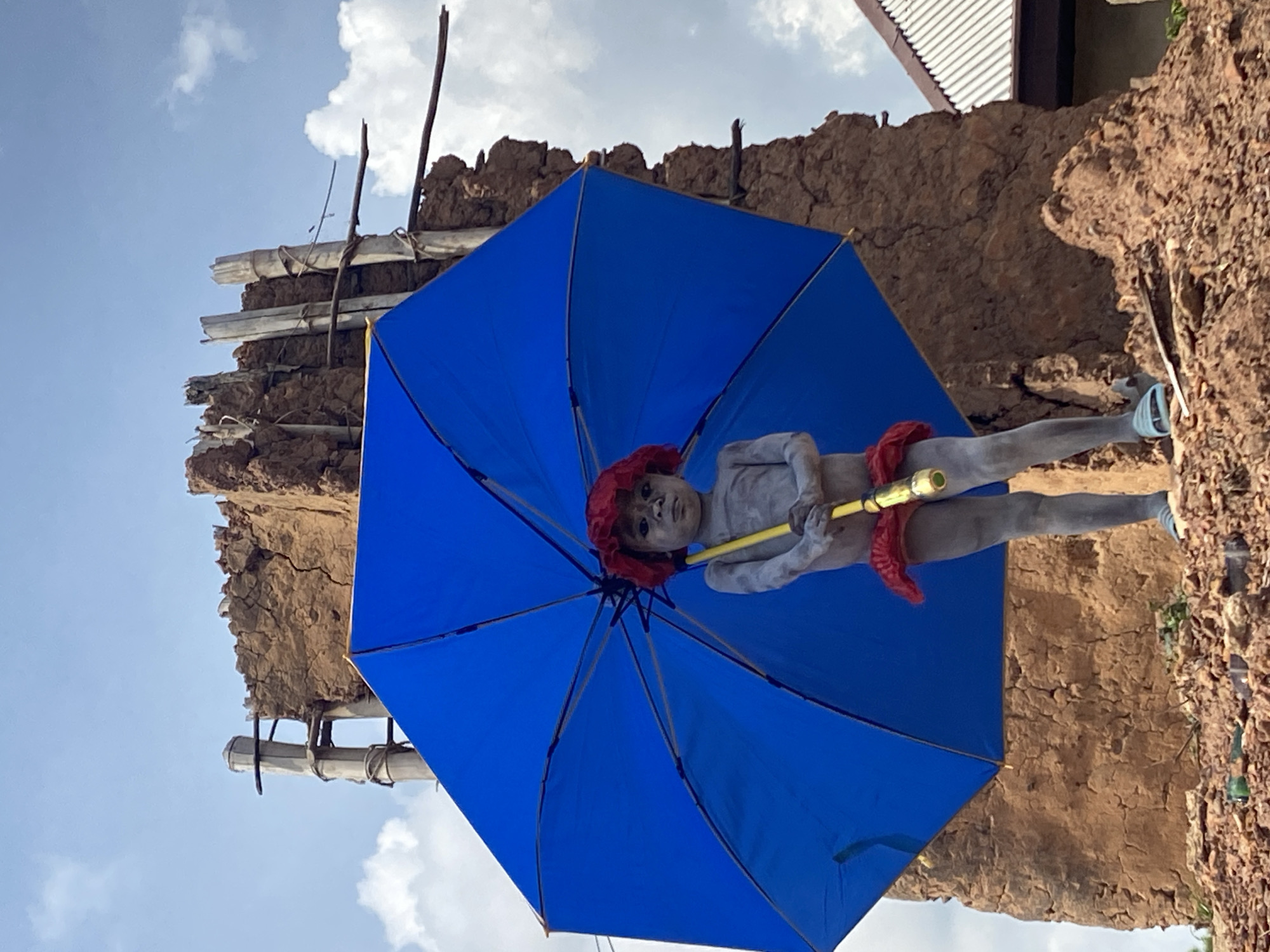 A person with gray body paint and a red headdress stands in front of a weathered, earthen structure under a large blue umbrella, likely at a cultural event or performance in Amoso, Nigeria. The earthen structure appears to be a building or wall with a deteriorating façade, suggesting an old or rural setting. The person is holding the umbrella's yellow handle, and red fabric is draped around their waist. The sky is visible in the background, with some clouds. The image is taken from a low angle, looking up at the person and the structure.