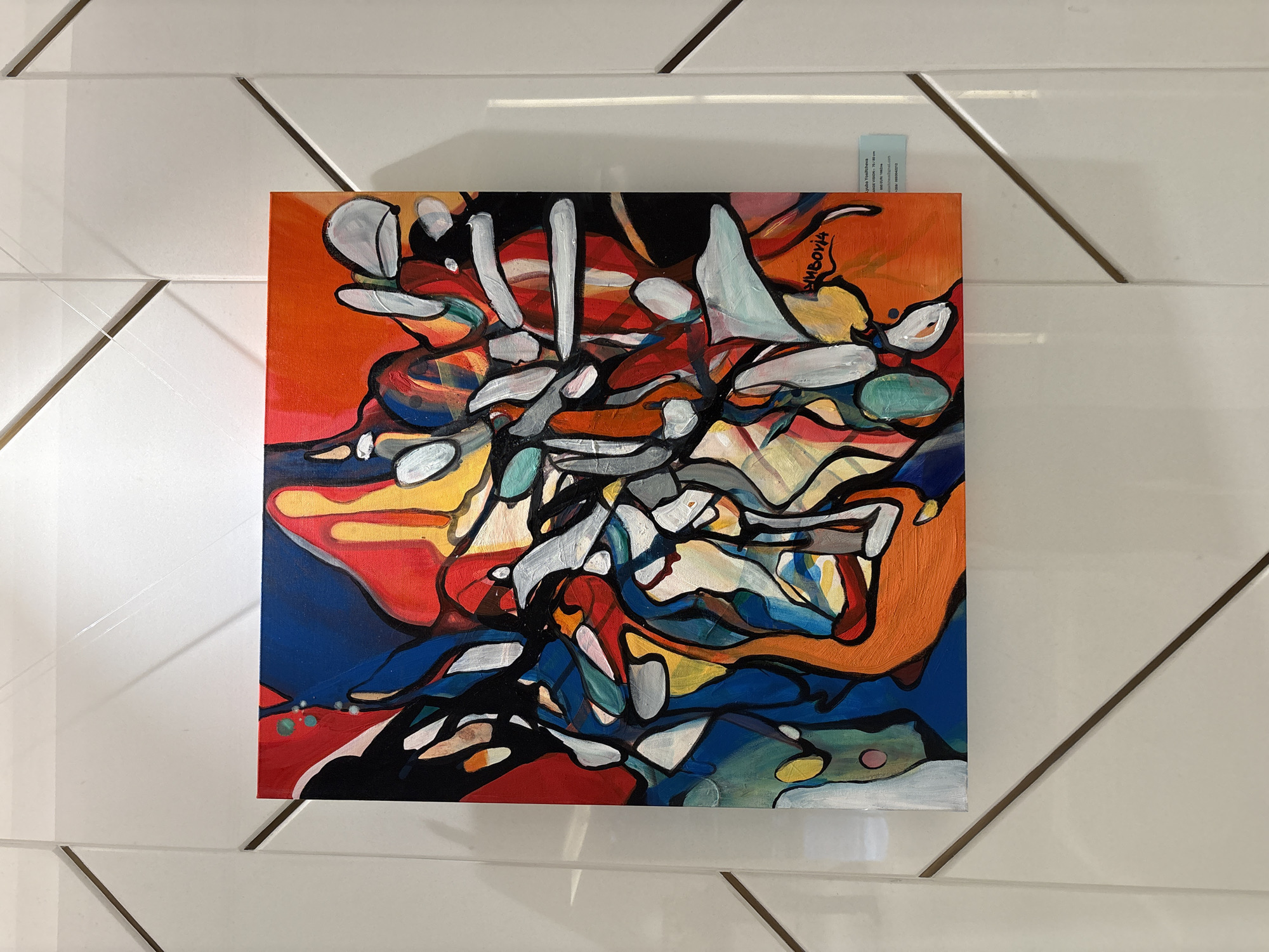 An abstract painting, square in shape and rich in color, is displayed against a white wall with a distinctive geometric pattern of diagonal lines. The painting features a dynamic composition of interconnected, organic, and angular shapes, primarily outlined in black. Its palette is vibrant, dominated by bold orange, deep red, and dark blue, with significant elements of bright yellow, white, and various shades of grey, along with accents of green and turquoise. The overall impression is one of chaotic energy and layered forms.
In the upper right corner of the painting, the word "KUMBOVIA" is vertically inscribed in a stylized black script, likely a signature or title. A small, light blue information card is tucked behind the top right edge of the artwork. Visible text on this card includes "Lyuba Vouchkova," "ART STUDIO - 70/80 cm," "vouchkova@gmail.com," and "tel: 0889242252." The "70/80 cm" likely refers to the dimensions of the artwork. The setting appears to be an indoor exhibition space, possibly in Sofia, Bulgaria, given the provided context.