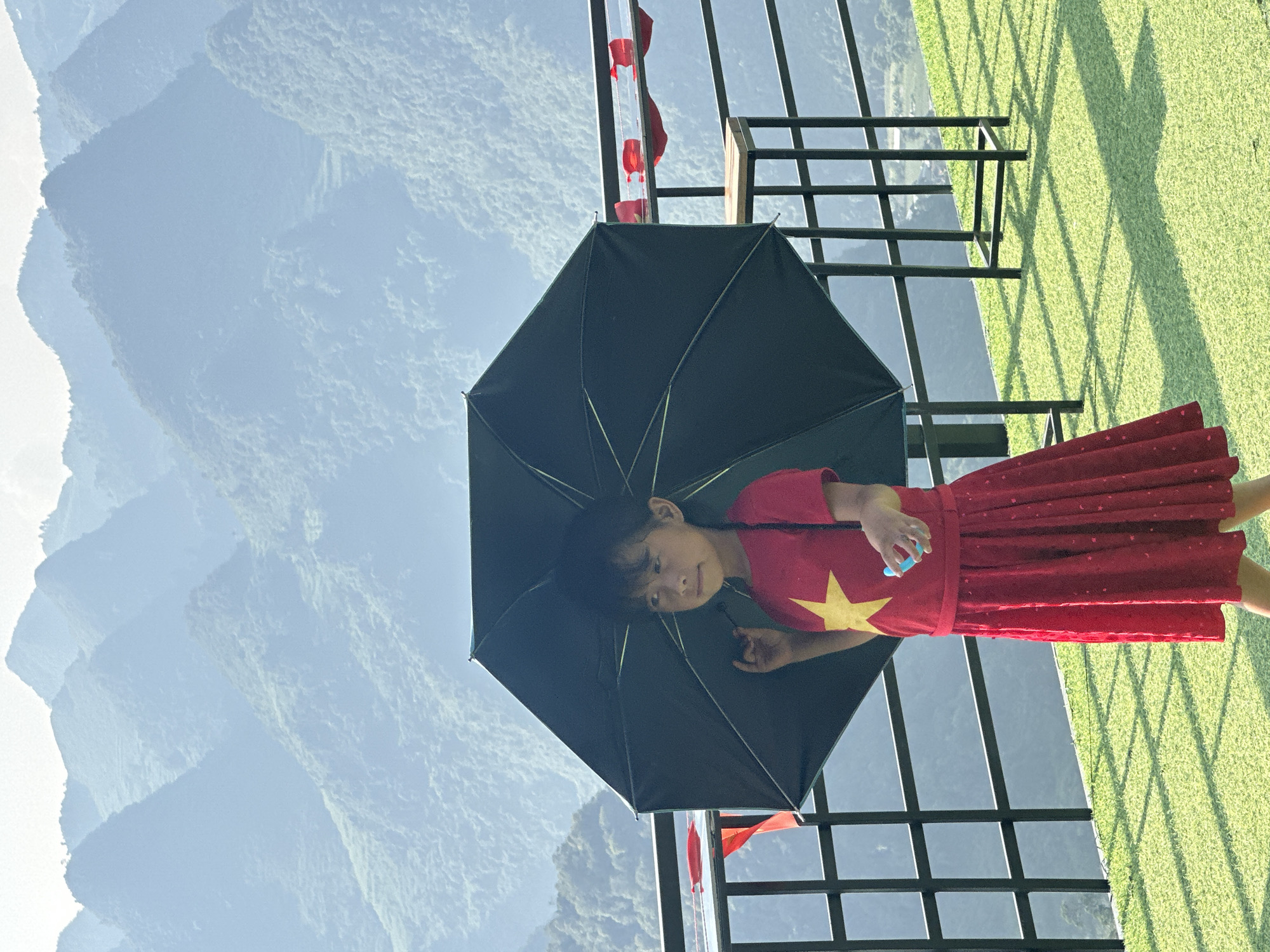 A young girl stands under a black umbrella in Đường Thượng Commune, Vietnam. She is wearing a red dress with a gold star, similar to the flag of Vietnam. The background features lush green grass, a black metal railing, and a mountainous landscape.