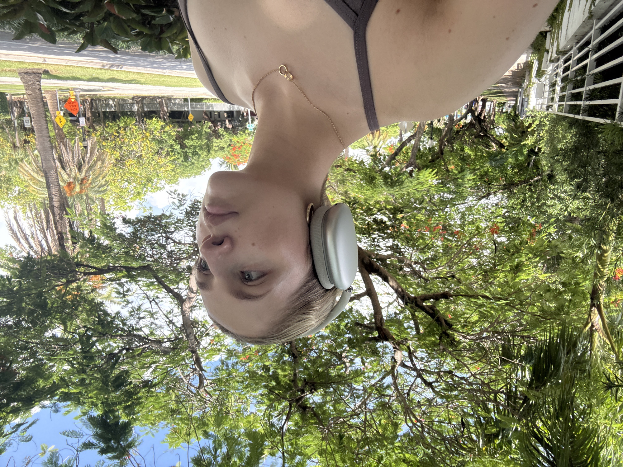 A young Caucasian woman with blonde hair pulled back is captured in a close-up selfie taken outdoors. She is wearing silver over-ear headphones, a grey tank top, and a delicate gold necklace with two intertwined rings. Her gaze is directed to her right, and her skin is lightly tanned with a few freckles visible on her shoulder.
The background suggests a vibrant, urban setting, likely in Miami, Florida, United States of America, characterized by lush green trees with vibrant orange flowers, and palm trees. A white fence lines a walkway on the left side of the frame, and a street with parked cars and road signs is visible on the right. The lighting indicates it is daytime, possibly late morning or early afternoon, with the sun casting gentle shadows. The overall scene conveys a sense of a leisurely outdoor activity, such as a walk or jog, with the woman enjoying music or a podcast.