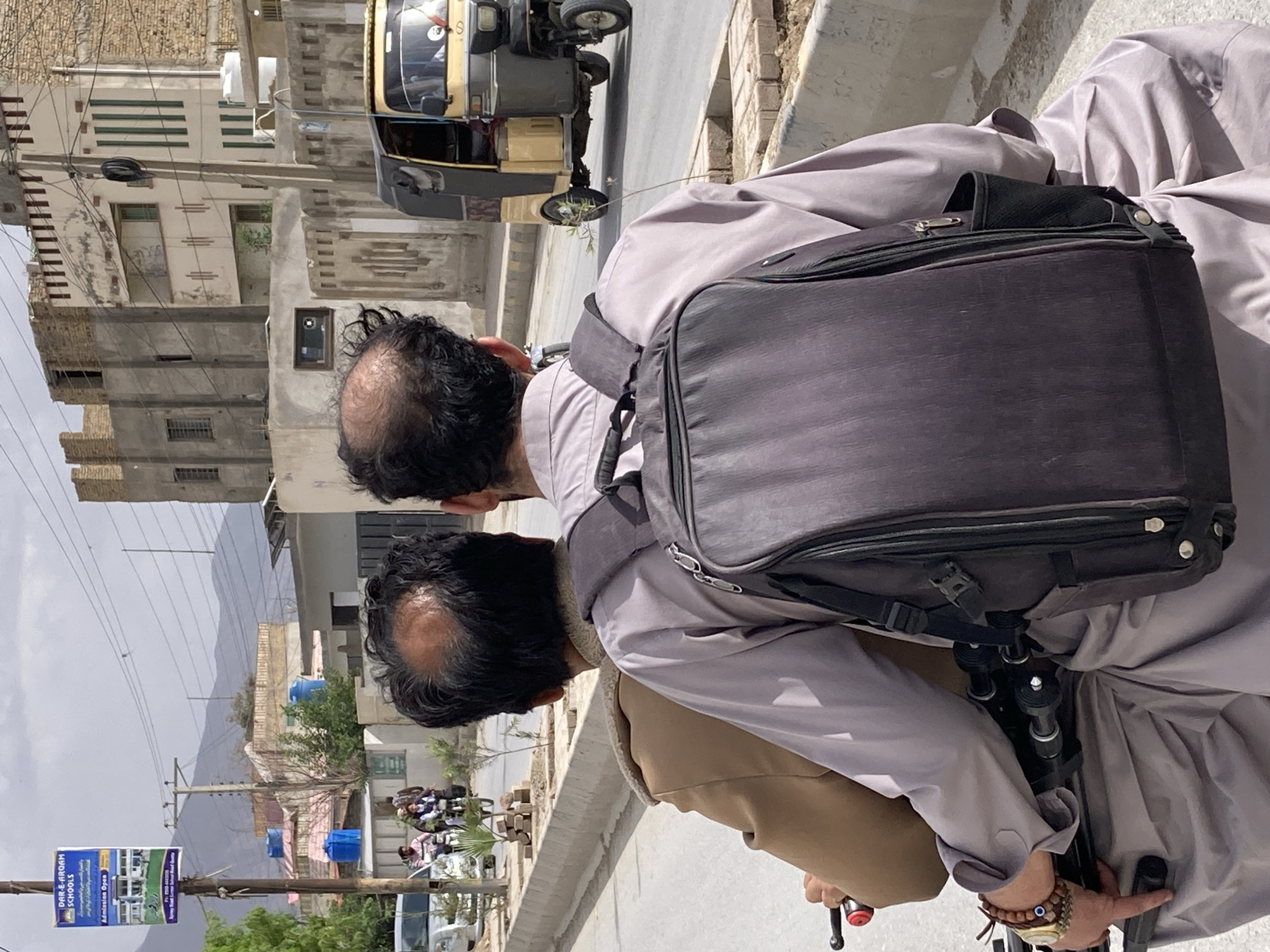 The image captures an outdoor street scene in Quetta City Tehsil, Pakistan, during daytime under an overcast sky. Two men are prominently featured in the foreground, seen from behind. Both have dark hair with visible balding on their crowns and are dressed in traditional light-colored attire, likely Shalwar Kameez. The man closer to the viewer also wears a tan-colored vest or jacket. Both are carrying dark backpacks; a camera tripod is attached to the backpack of the man in the immediate foreground. This man is also wearing a watch and a beaded bracelet with a blue evil eye charm on his right wrist, with his hand resting on a handlebar, suggesting they might be riding bicycles or a motorcycle, or are simply paused on the side of the road.
The paved road stretches into the background, flanked by various buildings, some appearing residential and others commercial, with signs of construction or weathering. In the mid-ground, a distinctive yellow and black auto-rickshaw is visible on the road. Further down the street, other vehicles, including a white car and what appear to be more motorcycles or bicycles, are present, along with groups of people. The area is characterized by numerous utility poles and tangled power lines. On one utility pole to the left, a blue banner is displayed, reading "DAR-E-ARQAM SCHOOLS" with "Admission Open" and contact details, along with images of school buildings. In the far background, majestic mountains rise under the cloudy sky, providing a natural boundary to the urban landscape.