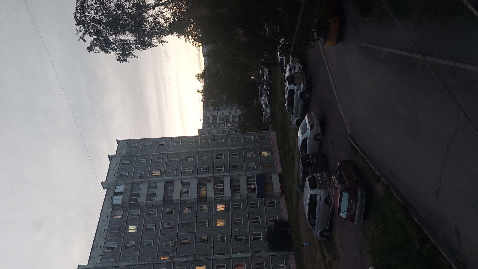 The image captures an outdoor scene in Amursk, Russia, at dusk. A large, multi-story apartment building, common in Soviet-era urban planning, dominates the left side of the frame. Its facade is a dull gray, with numerous windows, some of which are lit from within, suggesting occupancy. A balcony with glass enclosures is visible on one of the lower floors. In front of the building, a parking lot is filled with several cars. These include a silver minivan, a dark SUV, a white sedan, and a red station wagon parked closer to the foreground. The ground in the parking area appears to be a mixture of asphalt and gravel with patches of grass. To the right of the apartment building and parking lot, a dense cluster of dark trees partially obscures the sky and another apartment building in the distance. The sky is overcast with a pale, hazy glow near the horizon, indicating the setting sun. A thin wire stretches diagonally across the upper portion of the image. The overall atmosphere is subdued, characteristic of early evening in a residential urban environment.