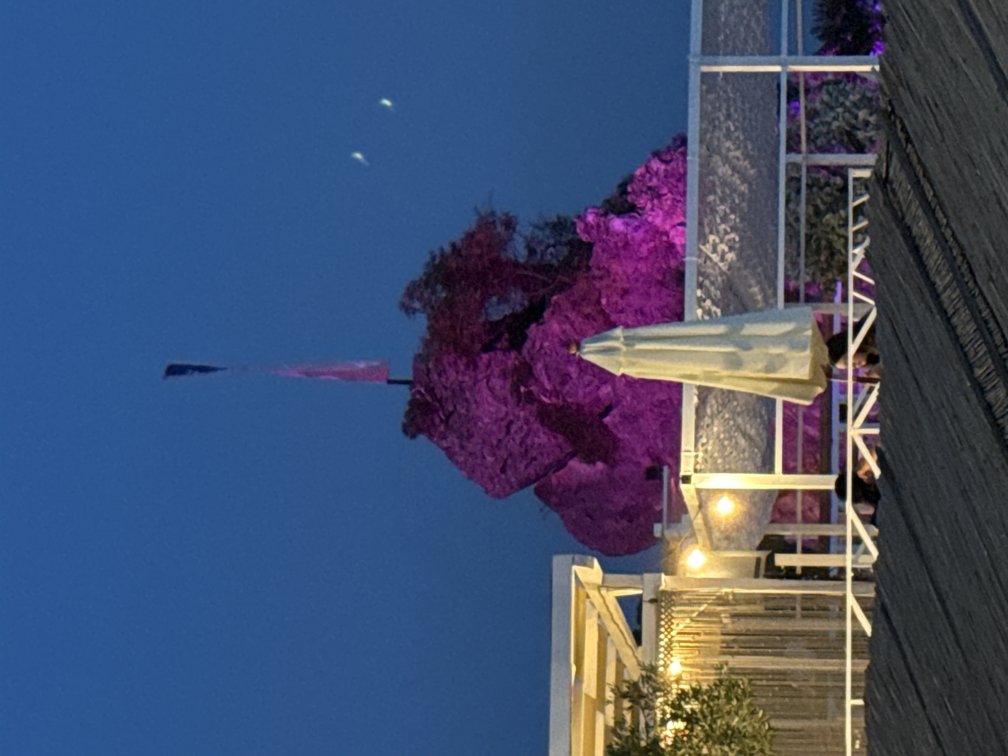 The image depicts a nighttime scene in Budva, Montenegro, featuring a rock formation illuminated with pink lights. A flag is visible at the summit of the rock. In the foreground, part of a thatched roof structure is present. Behind the rock, a white umbrella is visible, along with a white railing and part of a building with illuminated lights. The sky is dark blue, and two faint bright spots are present in the upper right quadrant, possibly distant lights or celestial bodies. A person is partially visible behind the railing. The overall ambiance suggests an outdoor dining or entertainment area.