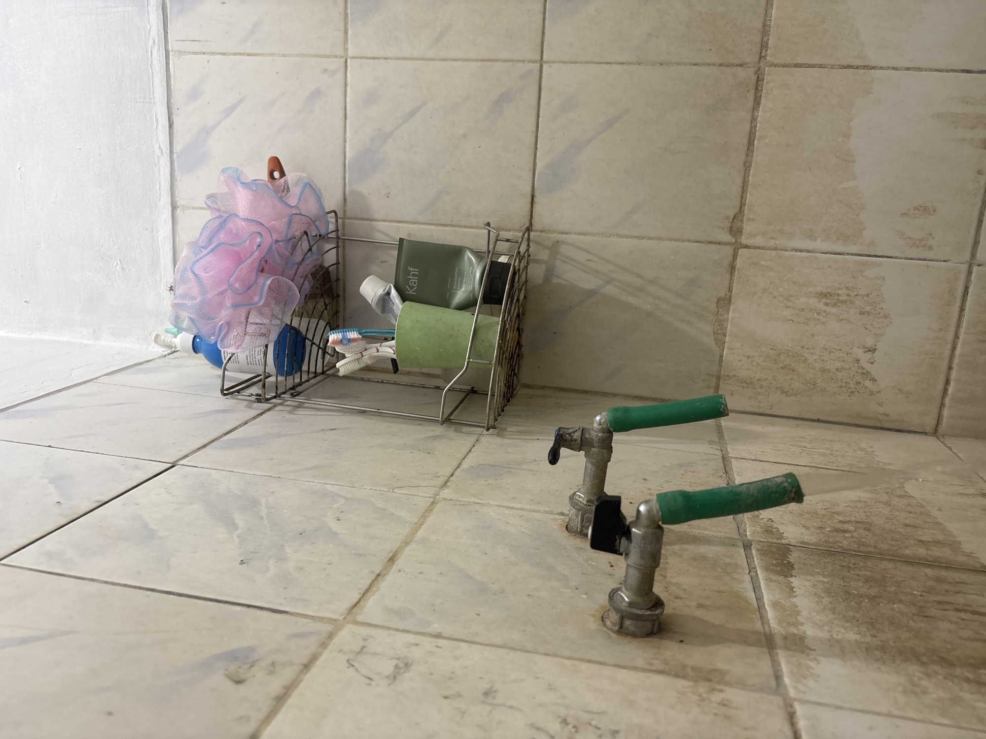 The image depicts a corner of a bathroom, likely in Palimanan, Indonesia, characterized by tiled walls and floors. Two metal faucets with green hose attachments are visible on the left side of the frame, with water flowing from one of them. The tiles appear aged and show signs of wear and staining, particularly around the faucets and on the lower portion of the wall.
Mounted on the wall above the faucets is a two-tiered wire rack containing various bathroom items. The top shelf holds a blue pump bottle and a large, pink loofah. The bottom shelf contains a green cup with toothbrushes and toothpaste, and a tube of "Kahf" brand skincare product. The overall impression is of a functional, albeit somewhat unkempt, bathroom space.