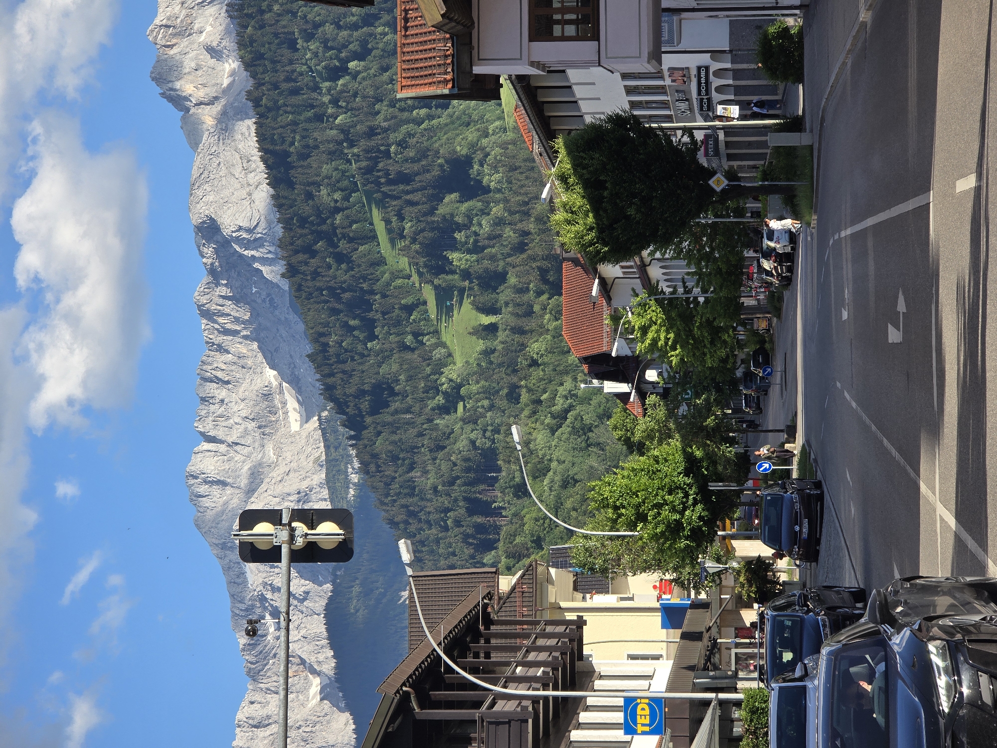 The image shows a street scene in Garmisch-Partenkirchen, Germany, with a backdrop of the Alps. In the foreground, a black Audi is parked on the left side of the road. Further down the street, traffic lights are visible, and several cars are parked along the side. Buildings line both sides of the street, with shops and apartments. A ski slope is visible on the green, tree-covered mountains in the distance. The sky is blue with scattered clouds.
