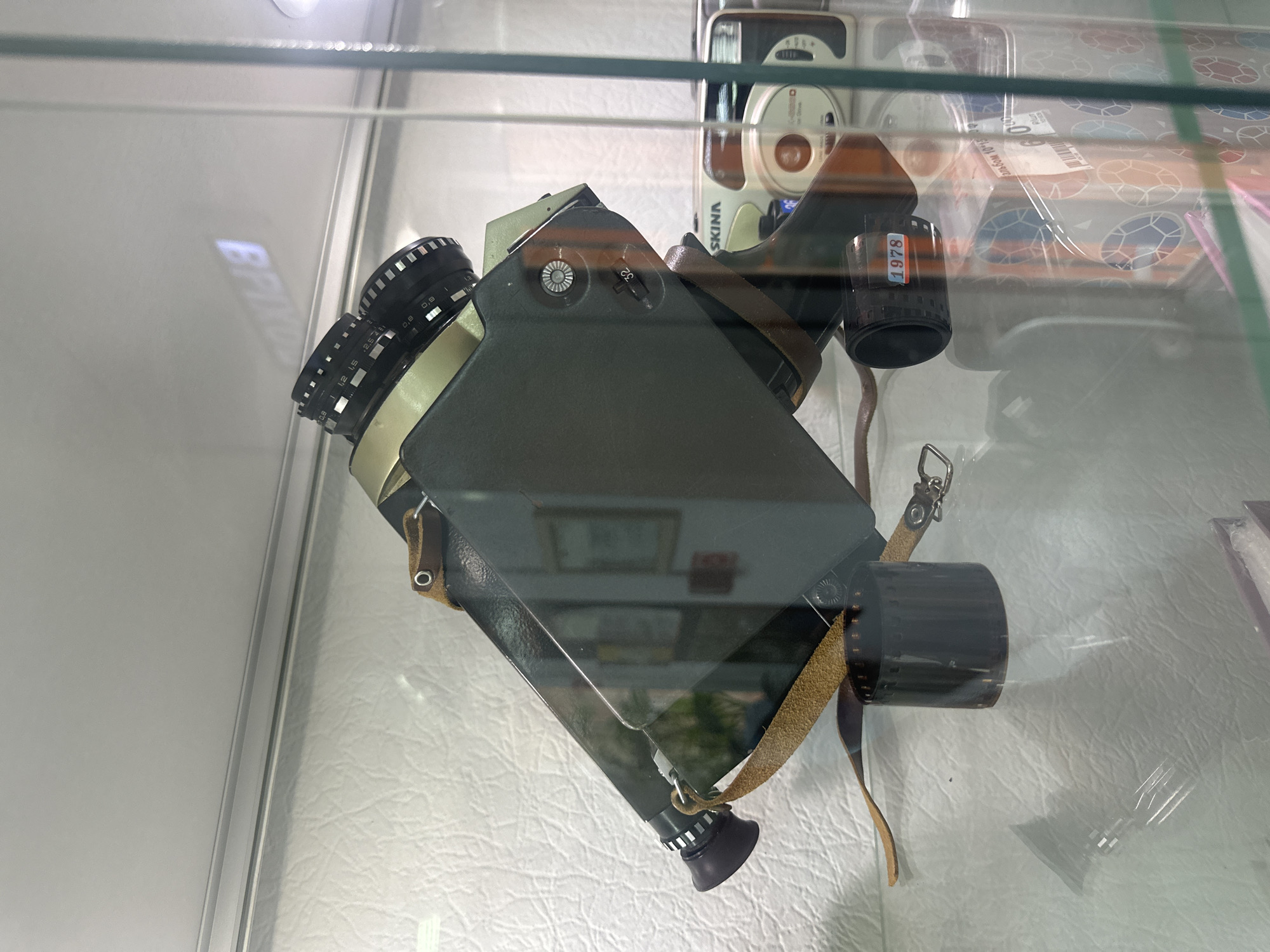 A vintage Super 8 film camera is displayed in a glass cabinet, angled upwards. The camera is dark gray with a gold-colored lens barrel and aperture rings. A brown leather strap is attached to the camera body and extends downwards. A roll of 8mm film is placed next to the camera, with a label reading "1978" on it. In the background, another vintage camera and a box with a colorful geometric pattern are visible. The background wall is textured white. The scene appears to be in a shop or museum display, and no people are present. The overall lighting is bright, creating reflections on the glass. There is a faint reflection of the word "BRIXTON" on the ceiling of the cabinet.