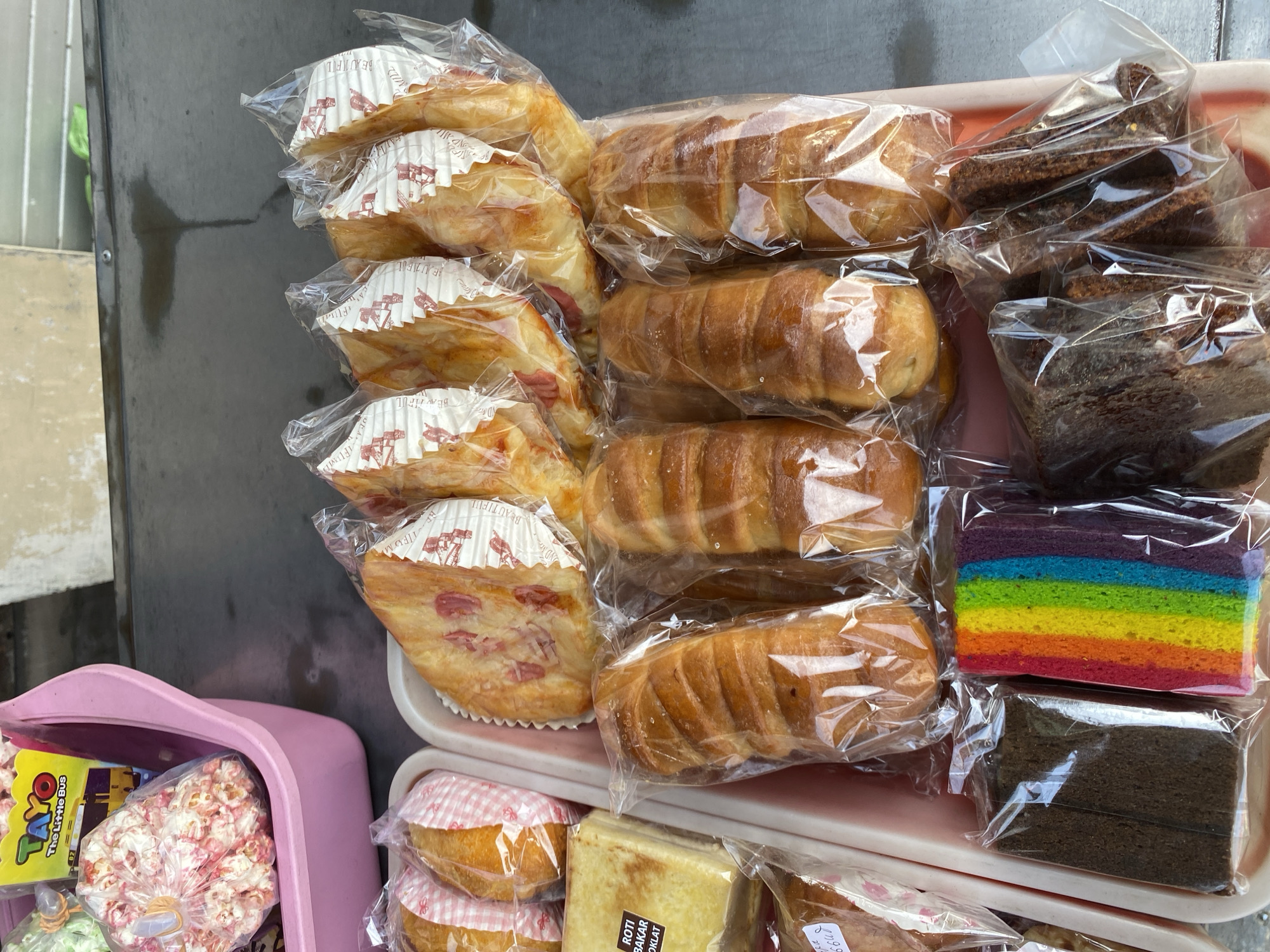 In Calukan RT 1 RW 5 Keboansikep, Indonesia, a display of baked goods is presented outdoors. In a pink plastic container on the left, there is popcorn, some of which is pink and white, and a yellow box labeled "TAYO The Little Bus". In the foreground, on a pink tray, are several types of bread and cakes, individually packaged in clear plastic. To the right of the popcorn, there are several pieces of bread with red toppings and white paper liners. Below these, there are several long, golden-brown loaves of bread, also individually wrapped. Further down and to the left are slices of cake, including a slice with rainbow-colored layers and a slice of dark chocolate cake. To the right of the rainbow cake, there are more dark brown slices of cake. The background shows a dark surface, possibly a table or counter, with a shadow.
