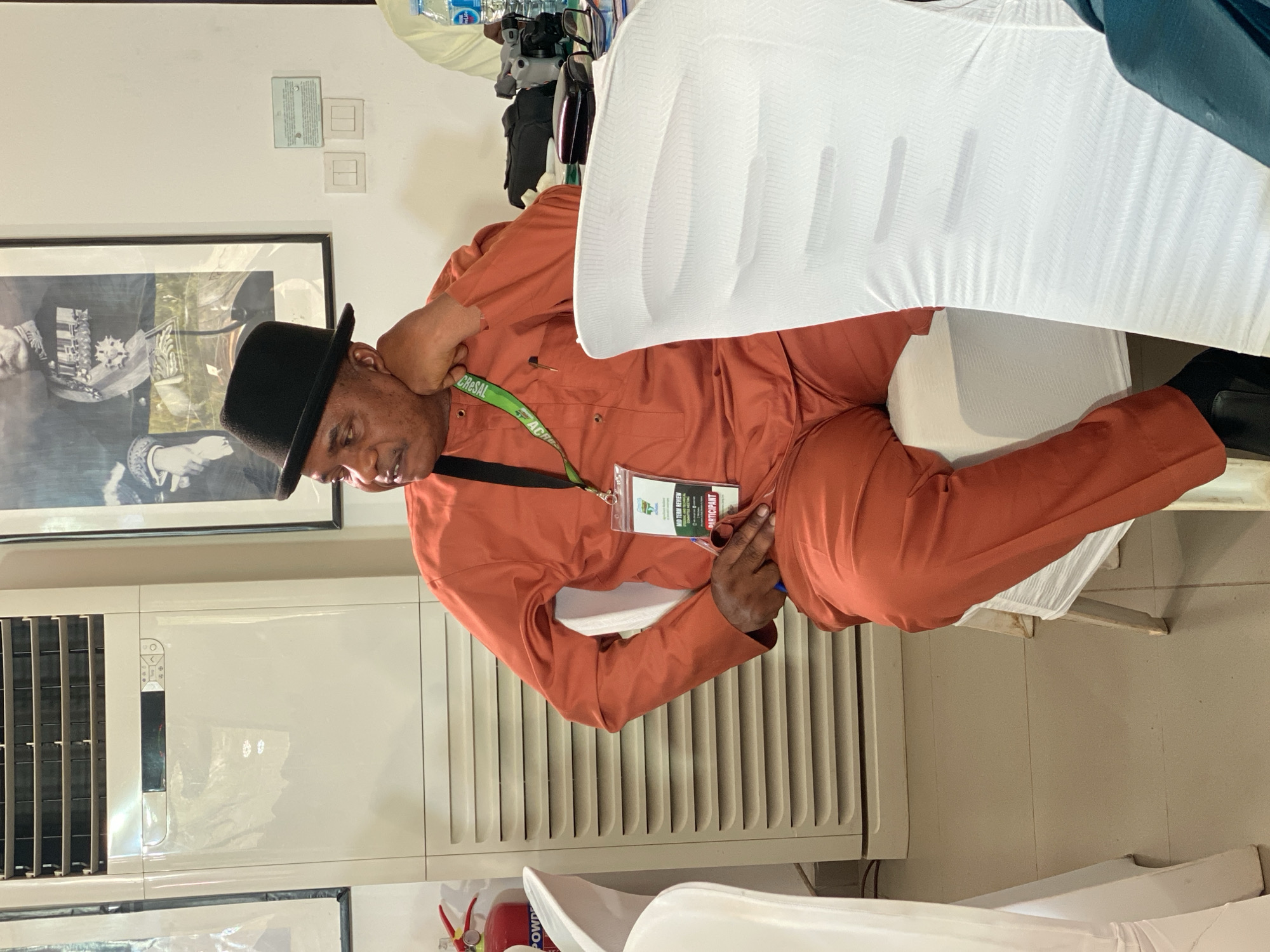 A medium, indoor shot depicts a man wearing a black hat and an orange jumpsuit. He is sitting in a white chair covered with a white cloth. He is wearing a lanyard with a name tag that reads "MID TERM REVIEW, PARTICIPANT." He is holding a pen in his right hand and resting his left hand on his neck. To his left is a white air conditioning unit and a framed photograph on the wall. A fire extinguisher is visible near the bottom left corner of the image. The location is presumed to be in Kwarau, Nigeria.