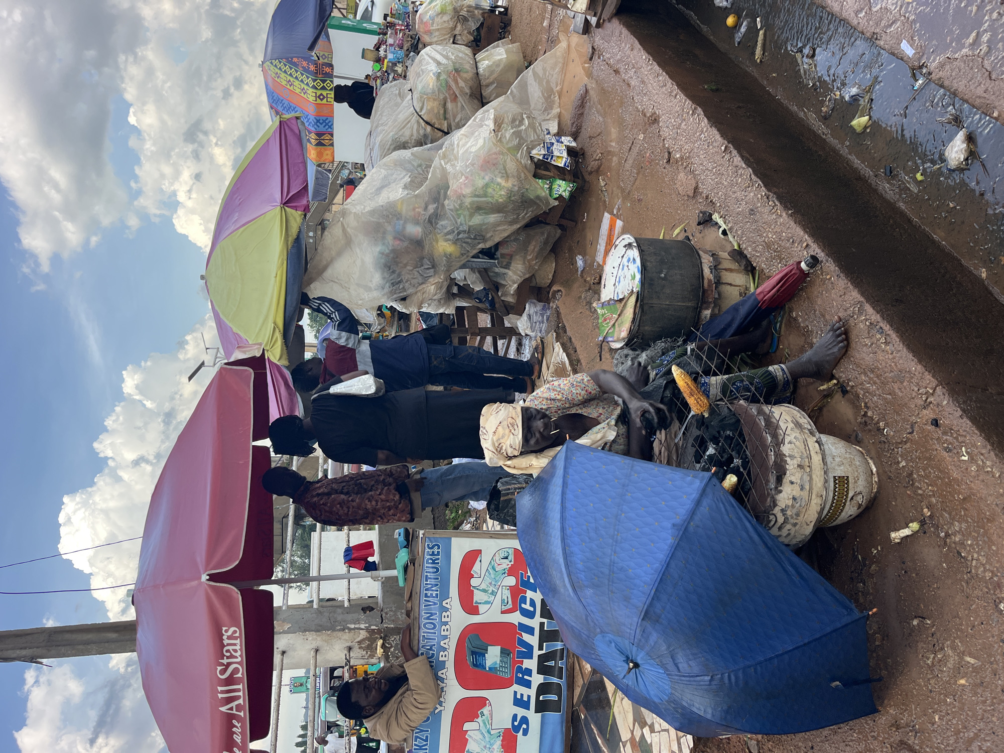 An outdoor scene depicts a busy marketplace with several people and stalls under various umbrellas. In the foreground, a woman sits on the ground next to a blue umbrella and a charcoal grill, preparing corn. She is wearing a headscarf and a patterned dress. Next to her, a man is sitting behind a counter that advertises "DOS SERVICE DATA" and "YAYA BABBA," with cash and a phone displayed on it. Further back, several people are standing and interacting, some near stalls selling goods wrapped in plastic. The ground is muddy and appears to be a street with a drainage ditch running along the side. The sky is cloudy.