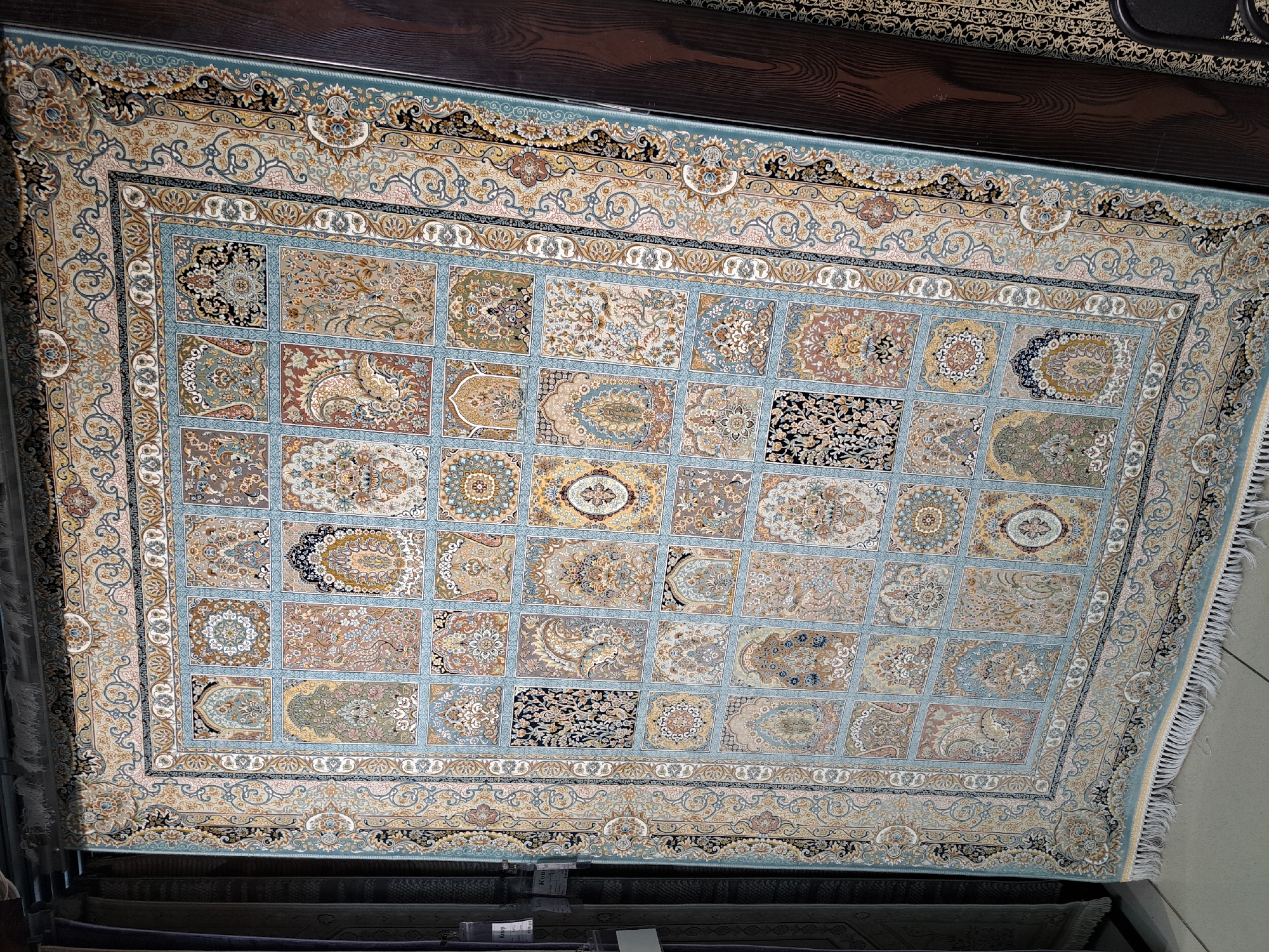 A large, intricately patterned Persian rug is displayed vertically in what appears to be a retail setting, possibly a carpet store or a market stall. The rug features a central panel divided into numerous smaller squares, each containing a unique floral or geometric design in shades of blue, beige, brown, and gold. The rug is bordered by ornate floral motifs and a fringe of white tassels at the bottom. The background shows a dark wooden surface and some other textiles, suggesting a commercial display. No people are visible in the image. The location is specified as Barnaul, Russia.