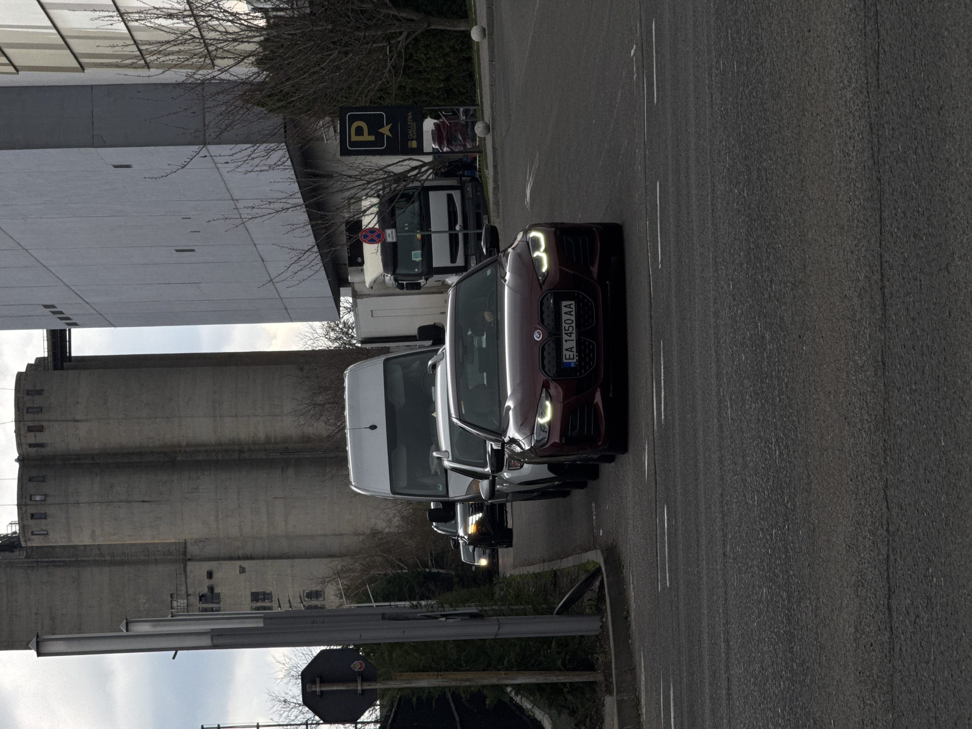 The image shows a street in Burgas, Bulgaria with several vehicles lined up. A burgundy BMW with license plate "EA 1450 AA" is in the foreground. Behind it, there is a white van and at least two other cars. The background includes a gray concrete structure, possibly a silo, as well as a modern white building. A parking sign with the letter "P" and the word "GALLERIA" is also visible. The street appears to be wet, and the lighting suggests it is either early morning or late afternoon.