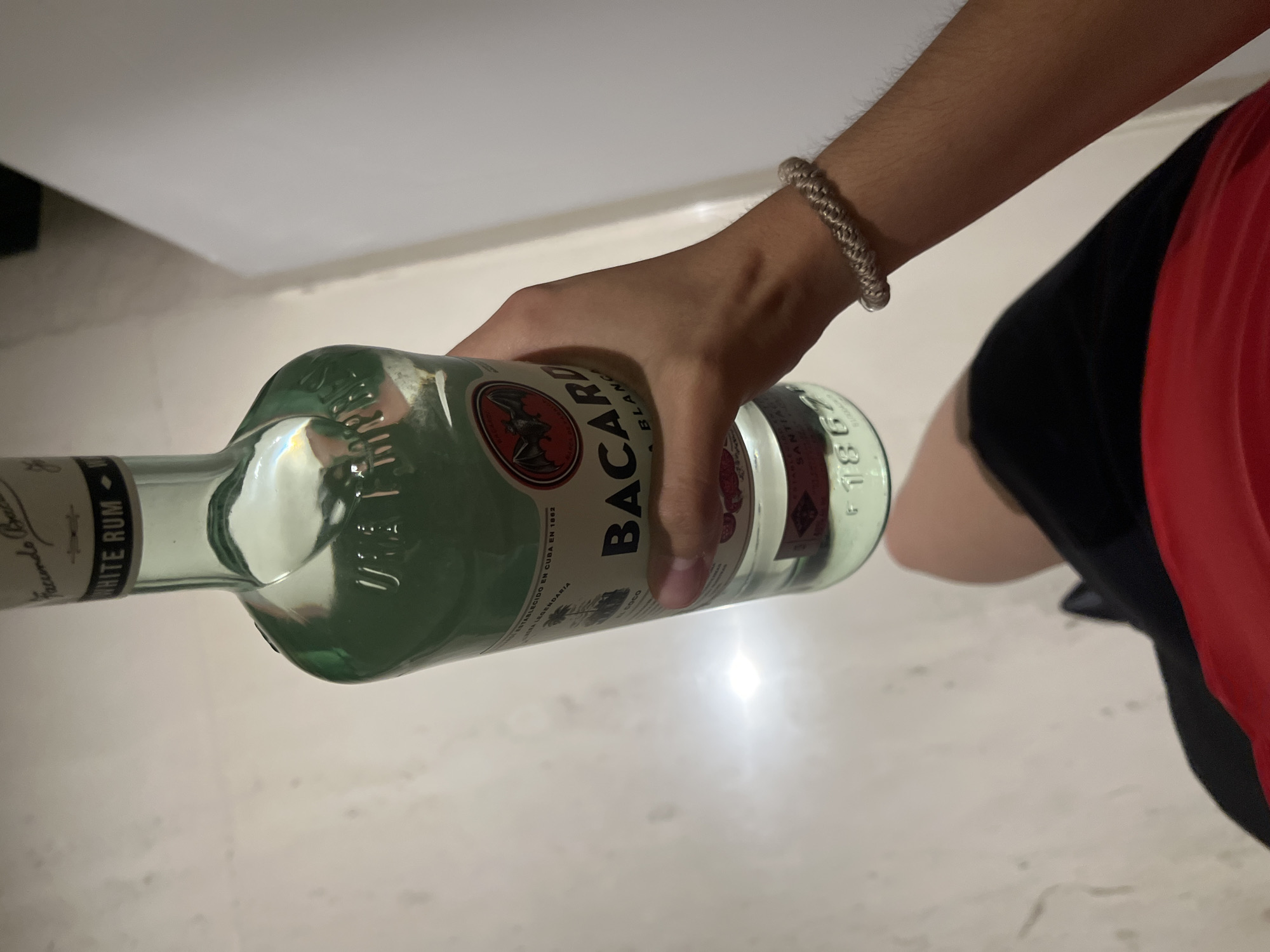 A person's right arm and upper torso are visible, holding a clear glass bottle of Bacardi White Rum horizontally. The hand firmly grips the bottle, which appears mostly full of clear liquid. The person's arm has light hair and wears a textured, light-colored bracelet on the wrist. They are dressed in a red top and black shorts.
The bottle's label features the text "BACARDI BLANC" and "WHITE RUM". Additional legible text includes "ESTABLECIDO EN CUBA EN 1862", "FACUNDO BACARDI", "LA LEGENDARIA", "COCO", "SANTIAGO", and "F1867". The numbers "81000A" are also visible.
The background consists of light-colored, possibly white or off-white, surfaces, indicating an indoor setting. Reflections are visible on the bottle's glass, suggesting artificial or diffused lighting. There are no direct visual cues within the image to determine the time of day or weather, nor any discernible emotions from the visible person. The provided location context for this media is San Bernardino Tlaxcalancingo, Mexico.