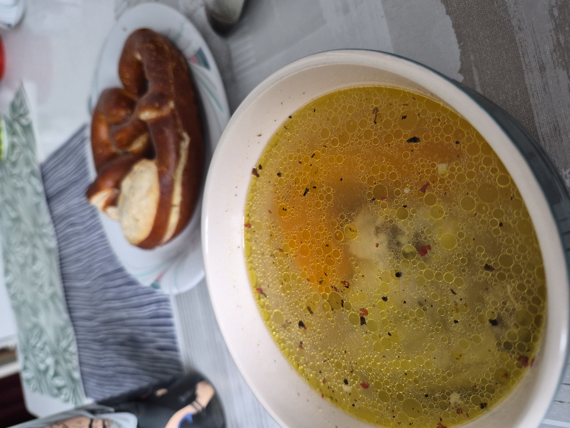 This is a top-down, close-up, slightly tilted shot of a meal on a table. The primary subjects are a bowl of soup and a pretzel on a plate. The soup is a light yellow broth with many small bubbles on the surface, indicating it is hot and perhaps freshly served. Scattered within the soup are small pieces of what appear to be herbs or spices, including some red flecks. A few larger chunks of food are visible beneath the surface. The pretzel is a classic shape, baked to a deep brown color, and appears to be fresh. It is placed on a white plate with a patterned rim. The table surface is a neutral gray, and a striped placemat or tablecloth is visible beneath the plate and to the left of the soup bowl. The setting appears to be indoors, likely a casual dining environment. There is no indication of time of day or weather. No people are visible in the frame, and no text is present. The overall impression is of a simple, hearty meal being presented.