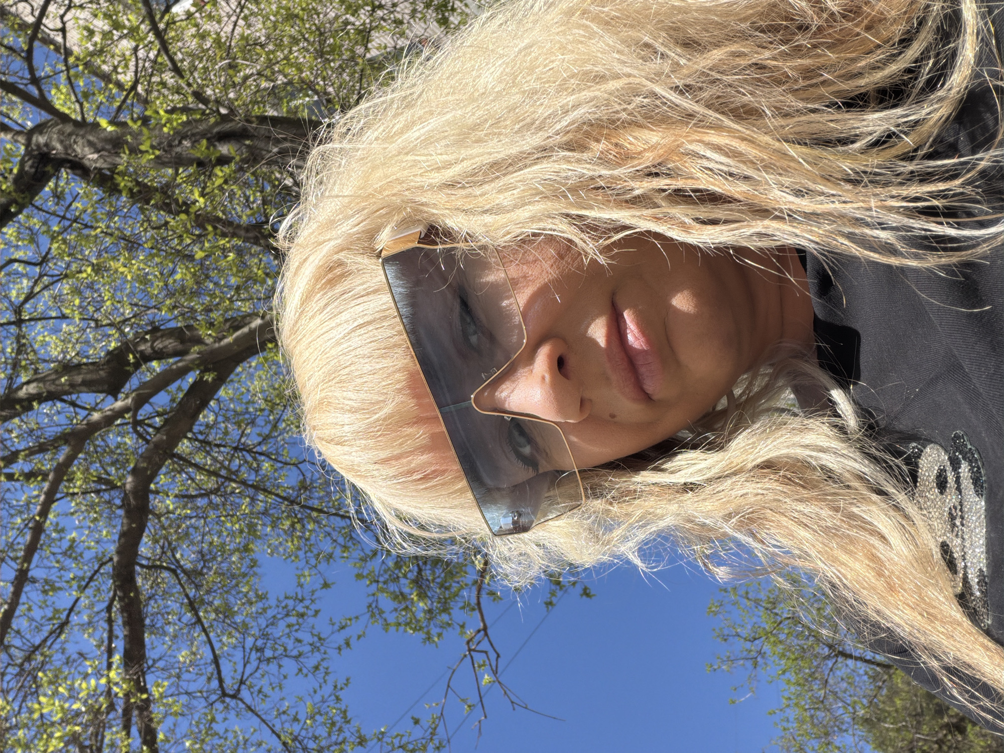A woman with blonde hair and futuristic sunglasses looks up at the camera. The background includes tree branches with green leaves against a bright blue sky. The woman is wearing a dark-colored top with a shiny silver design on the shoulder. The image was taken outdoors in daylight. I have no information that leads to this image being in Chaikovsky, Russia.