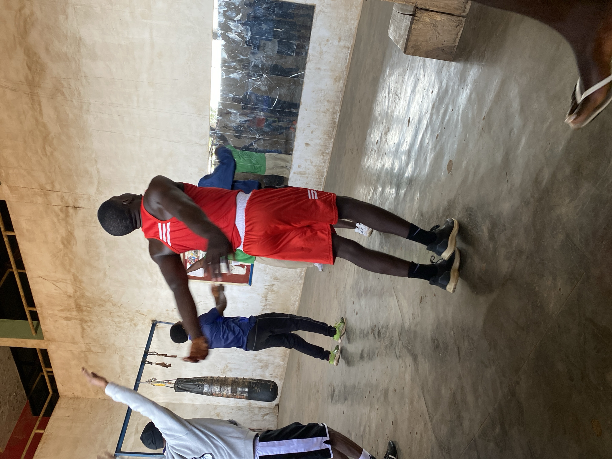 The image shows an indoor training space in Jos, Nigeria. A man in red boxing shorts and a matching tank top is practicing punches, facing away from the camera. To his left, a man in a blue shirt and black pants is also practicing punches. In the foreground, a third man wearing a white long-sleeved shirt and black shorts has one arm raised. A punching bag hangs from a metal frame in the background. The walls are tan, and there is a cloudy mirror on one wall. The floor is concrete.