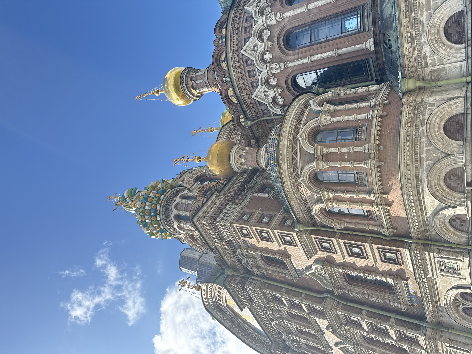 The image shows a low-angle view of the Church of the Savior on Spilled Blood in Saint Petersburg, Russia. The building's ornate facade, including its golden domes and intricate brickwork, is prominently displayed against a clear blue sky with a few scattered clouds. The architectural details showcase various windows and decorative elements.